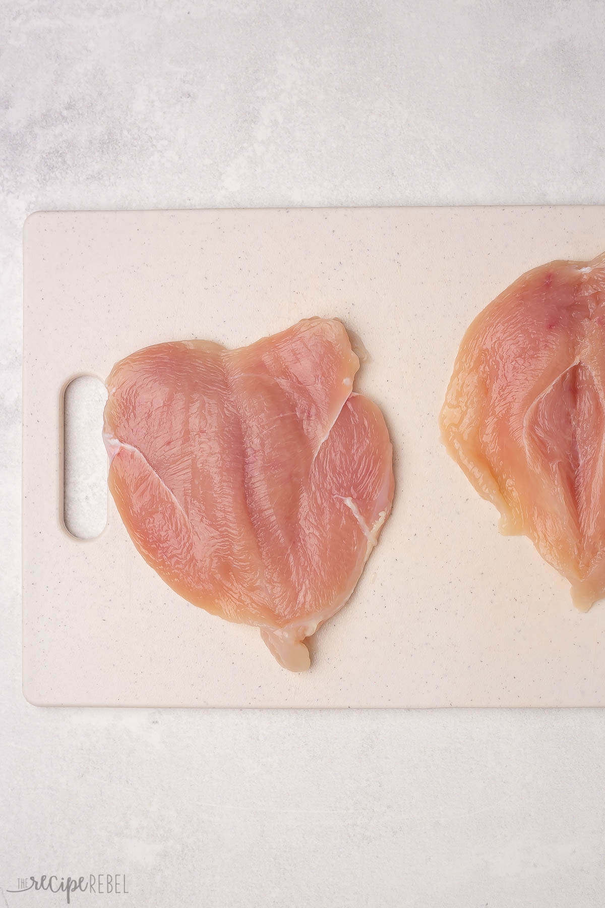 two chicken breasts sliced in half but not separated on white cutting board.