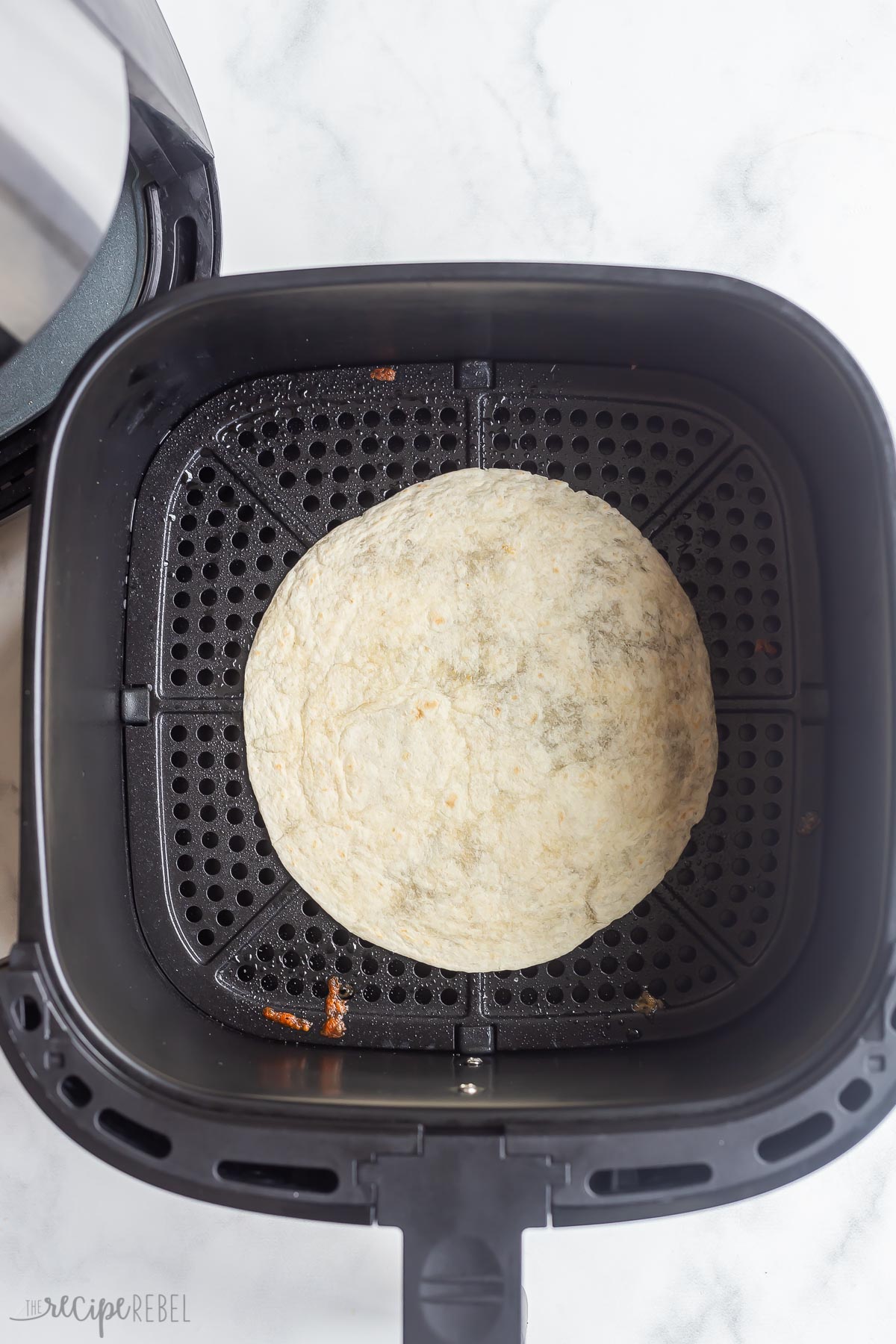 One tortilla laying flat in air fryer basket beside air fryer.