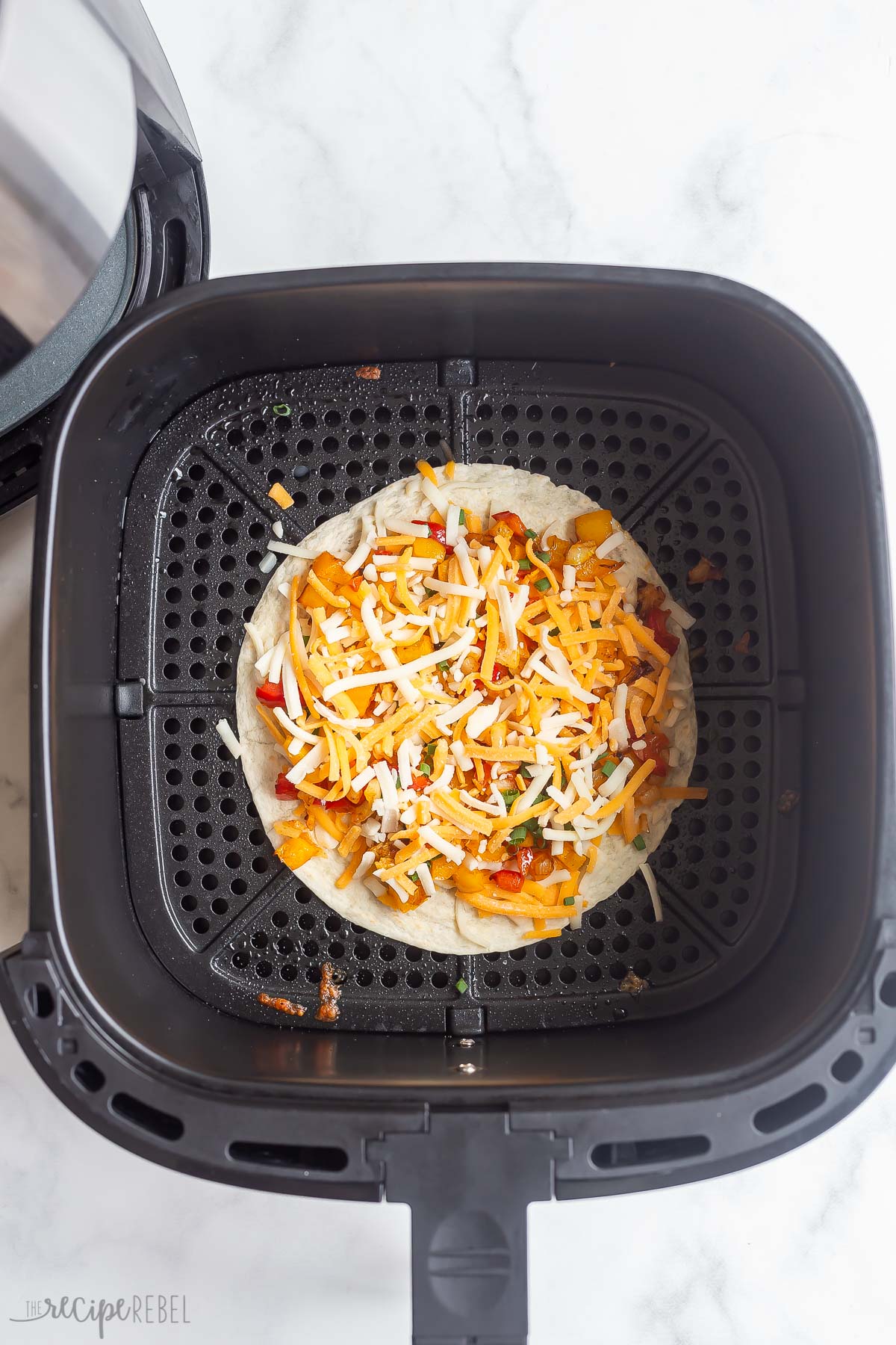 Top view of tortilla with toppings and shredded cheese in air fryer basket.