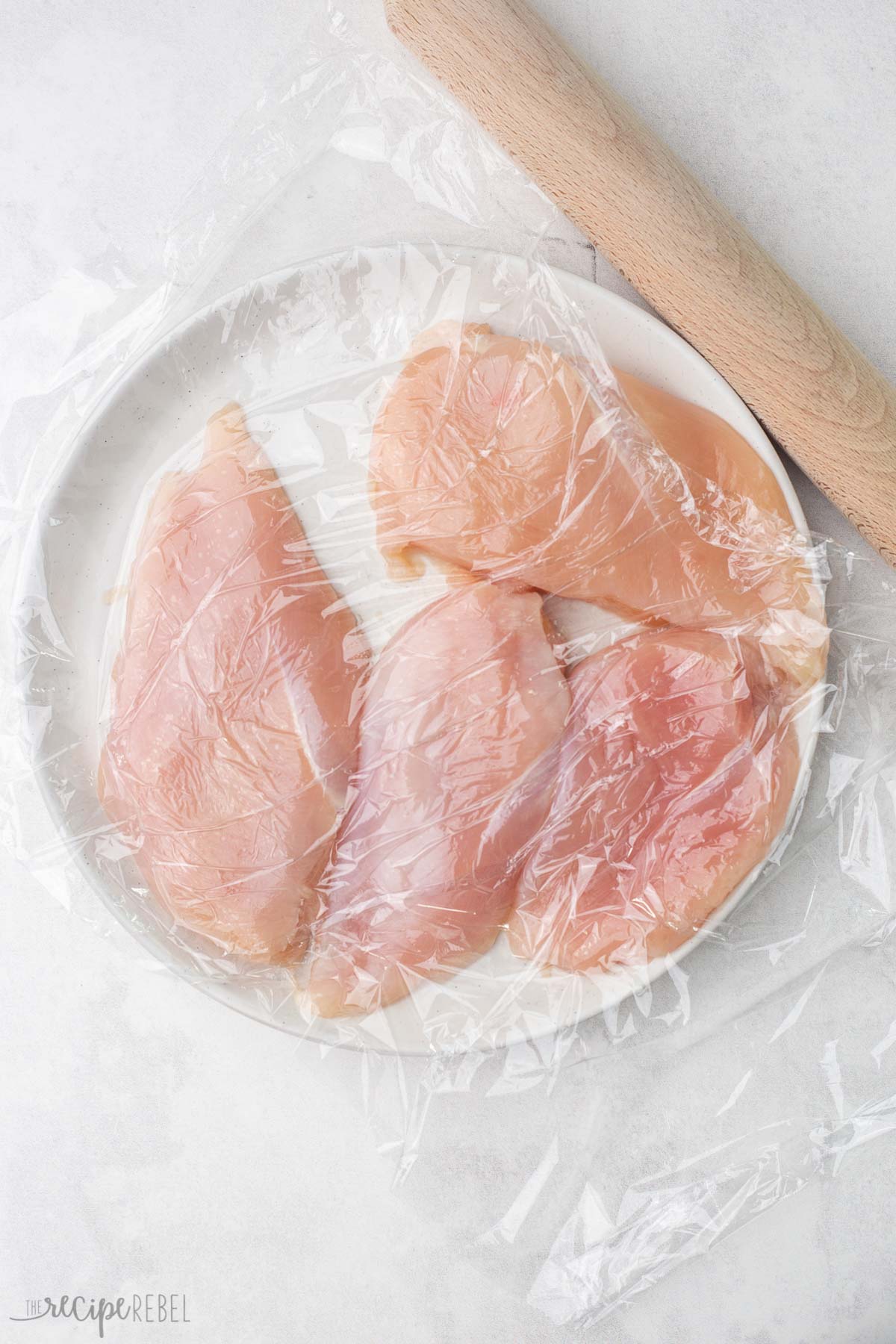 chicken breasts on a white plate covered in plastic wrap with rolling pin beside.