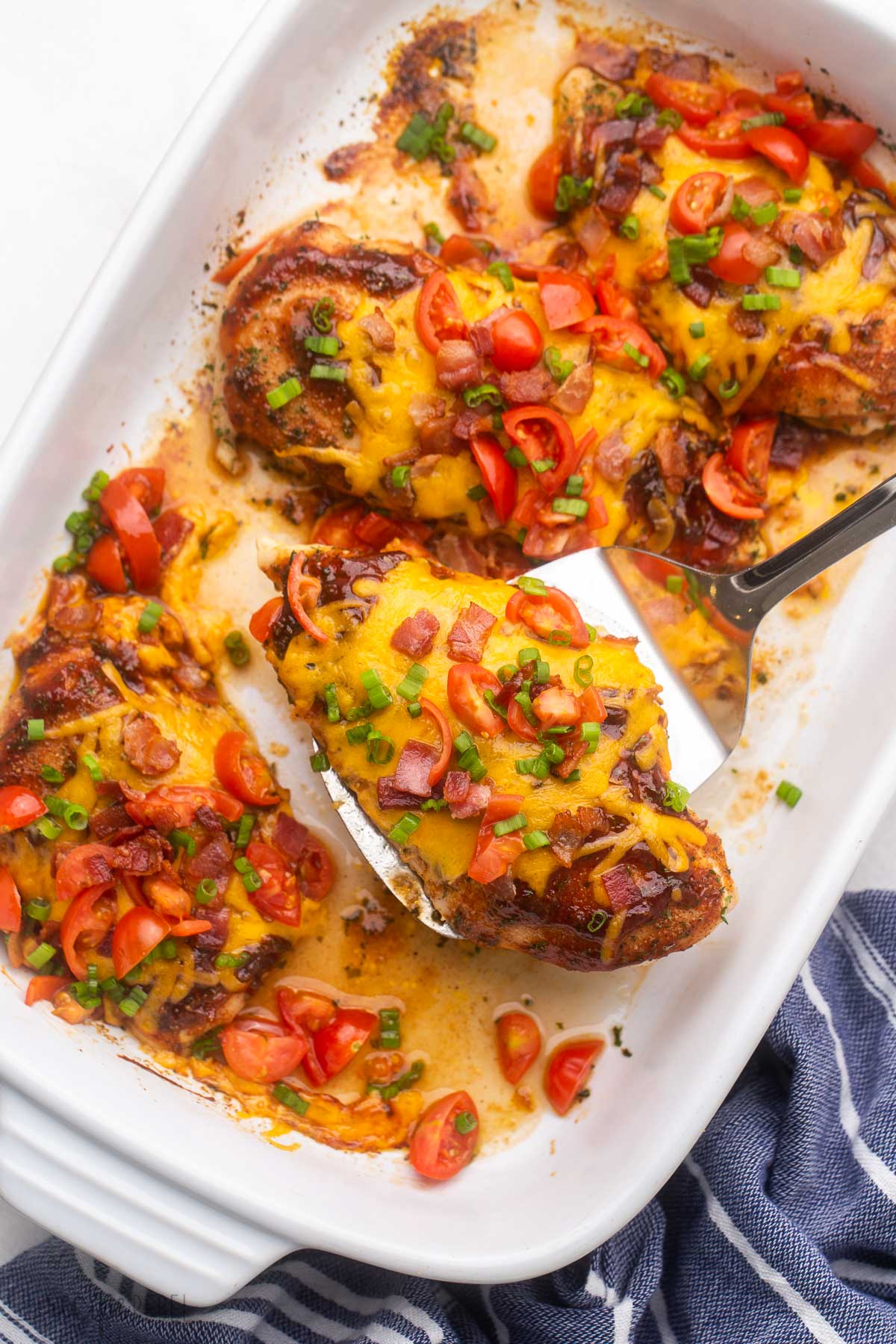 This Baked Monterey Chicken Recipe is so flavorful, with a combination of juicy baked chicken breasts, fresh tomatoes, Monterey Jack cheese, and a smoky bacon flavor from the first bite. #chicken #dinner | dinner ideas | dinner recipes | chicken breast | baked chicken | easy chicken recipe | bbq sauce overhead shot of spatula holding baked monterey chicken breast dish.