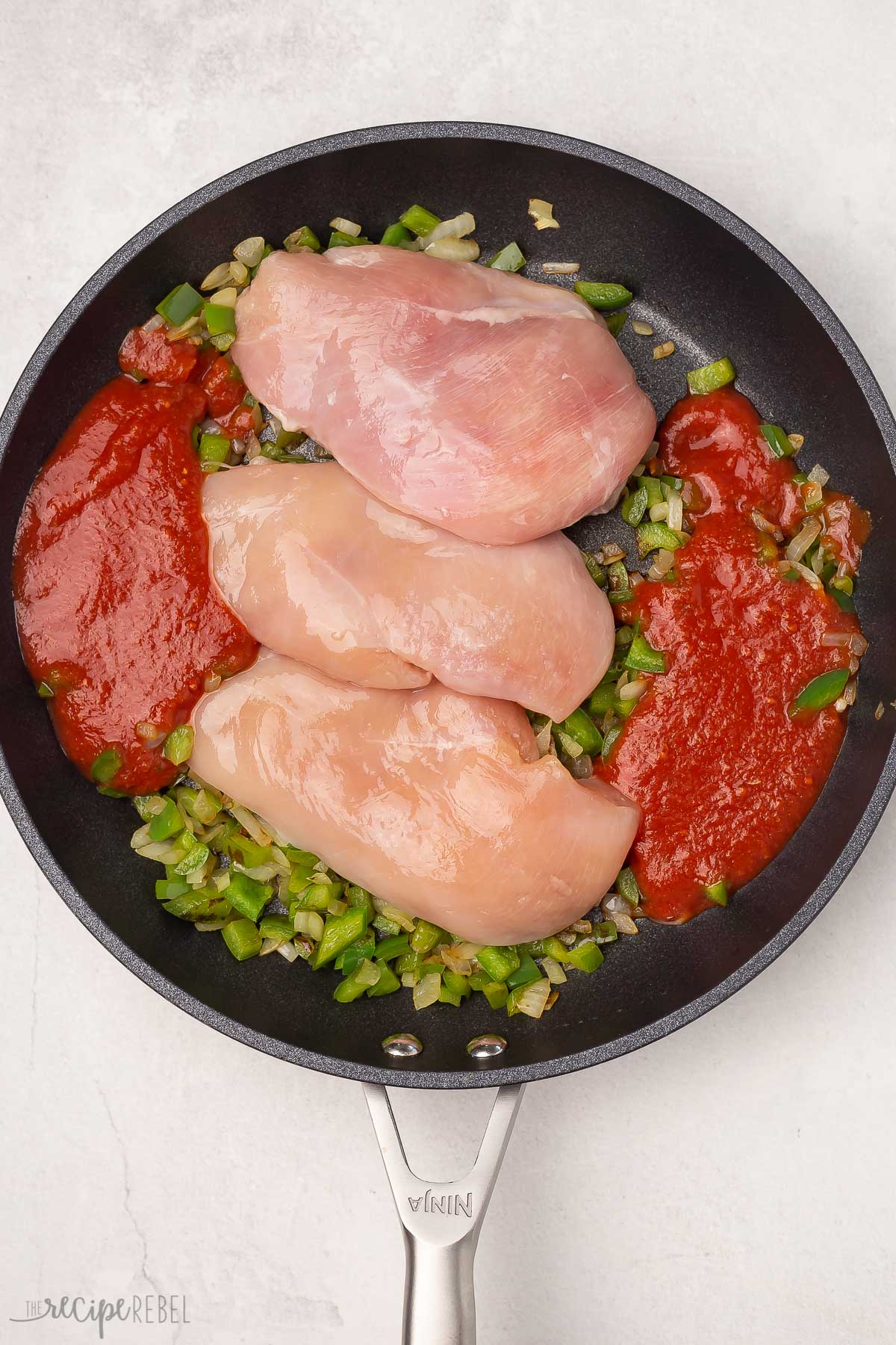 Top view of skillet with chopped onions, green bell peppers, red enchilada sauce, and three chicken breasts on top. 