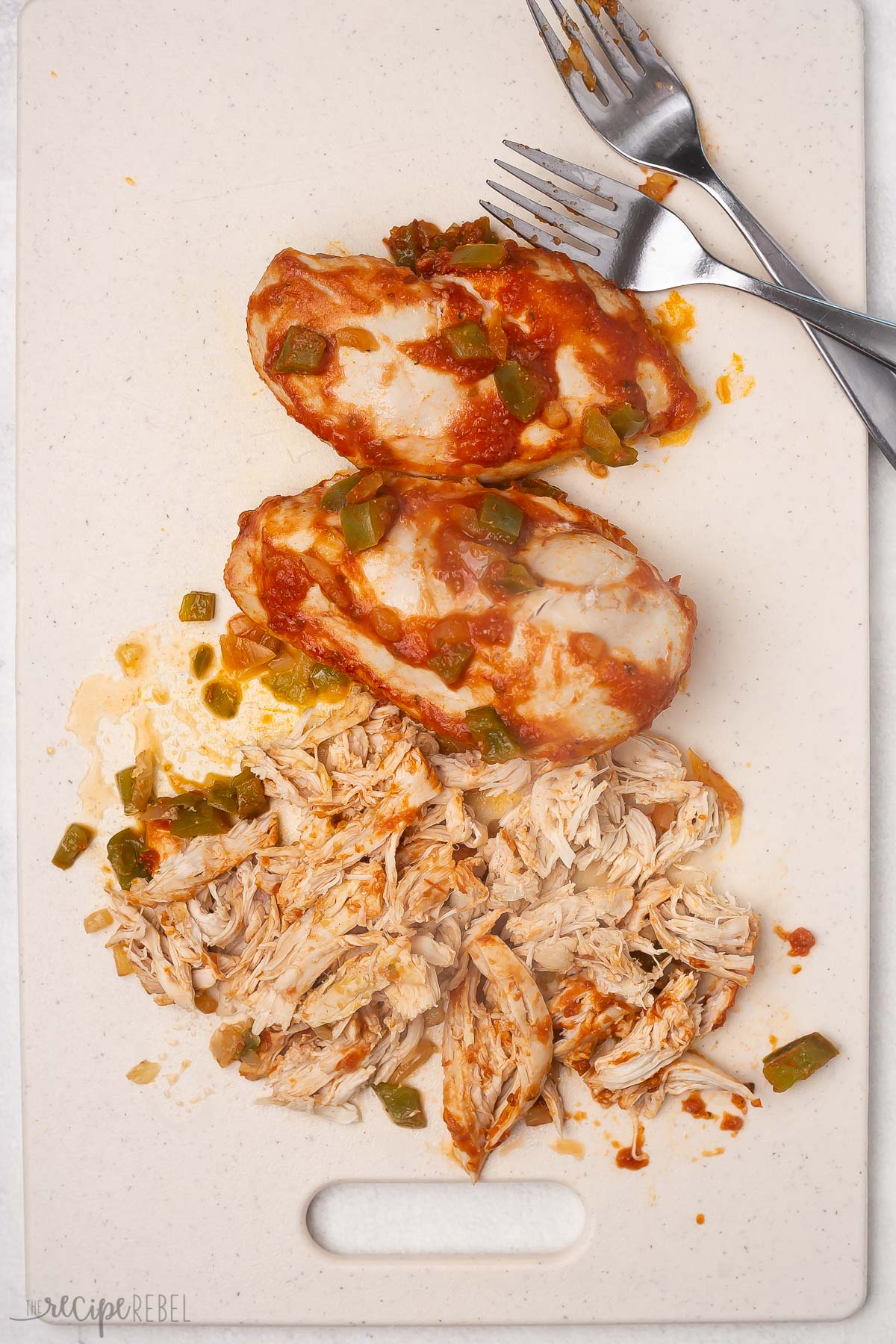 Top view of cooked chicken breasts with some red sauce on it being shredded on a chopping board with a fork. 