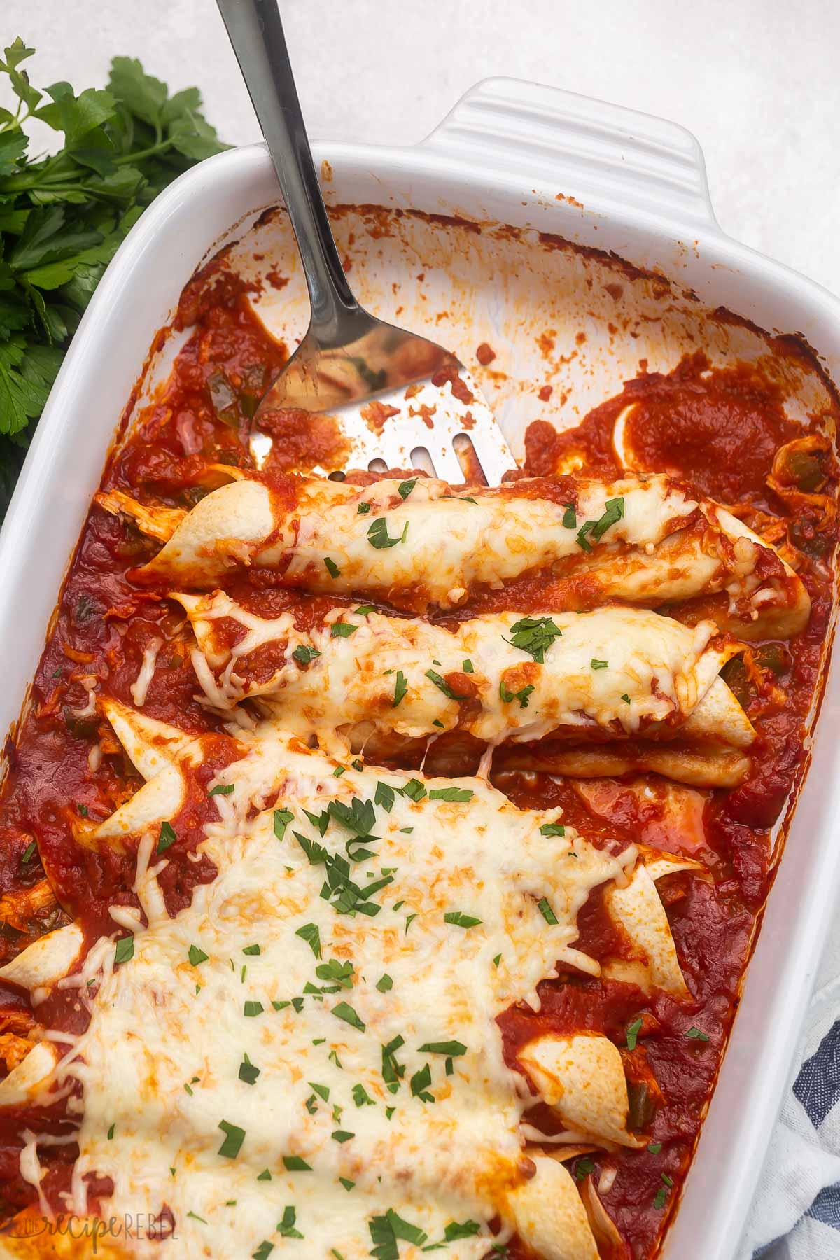 a white baking dish of enchiladas with a spatula.