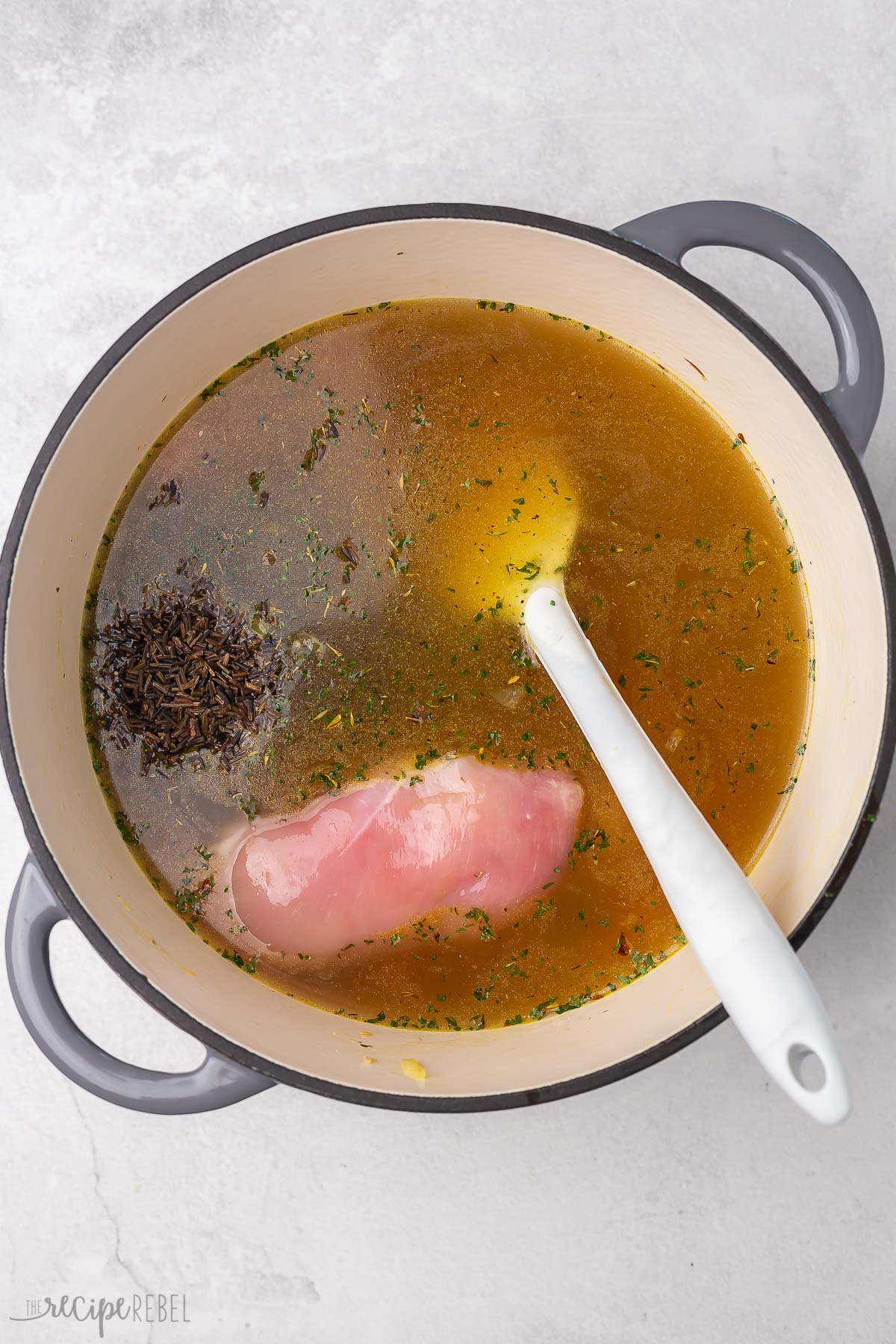 Top view of dutch oven with raw chicken breasts, chicken broth and seasoning being added on top of cooked vegetables. 