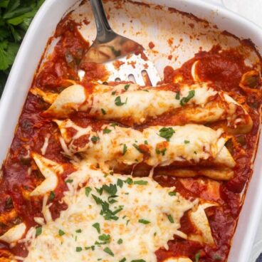 a white baking dish of enchiladas with a spatula.