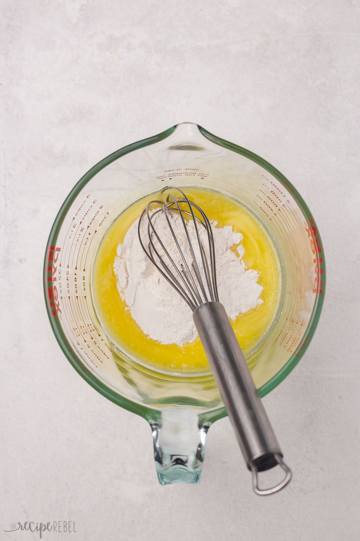 Top view of a glass jug with melted butter in the bottom and flour on top and a whisk in it.