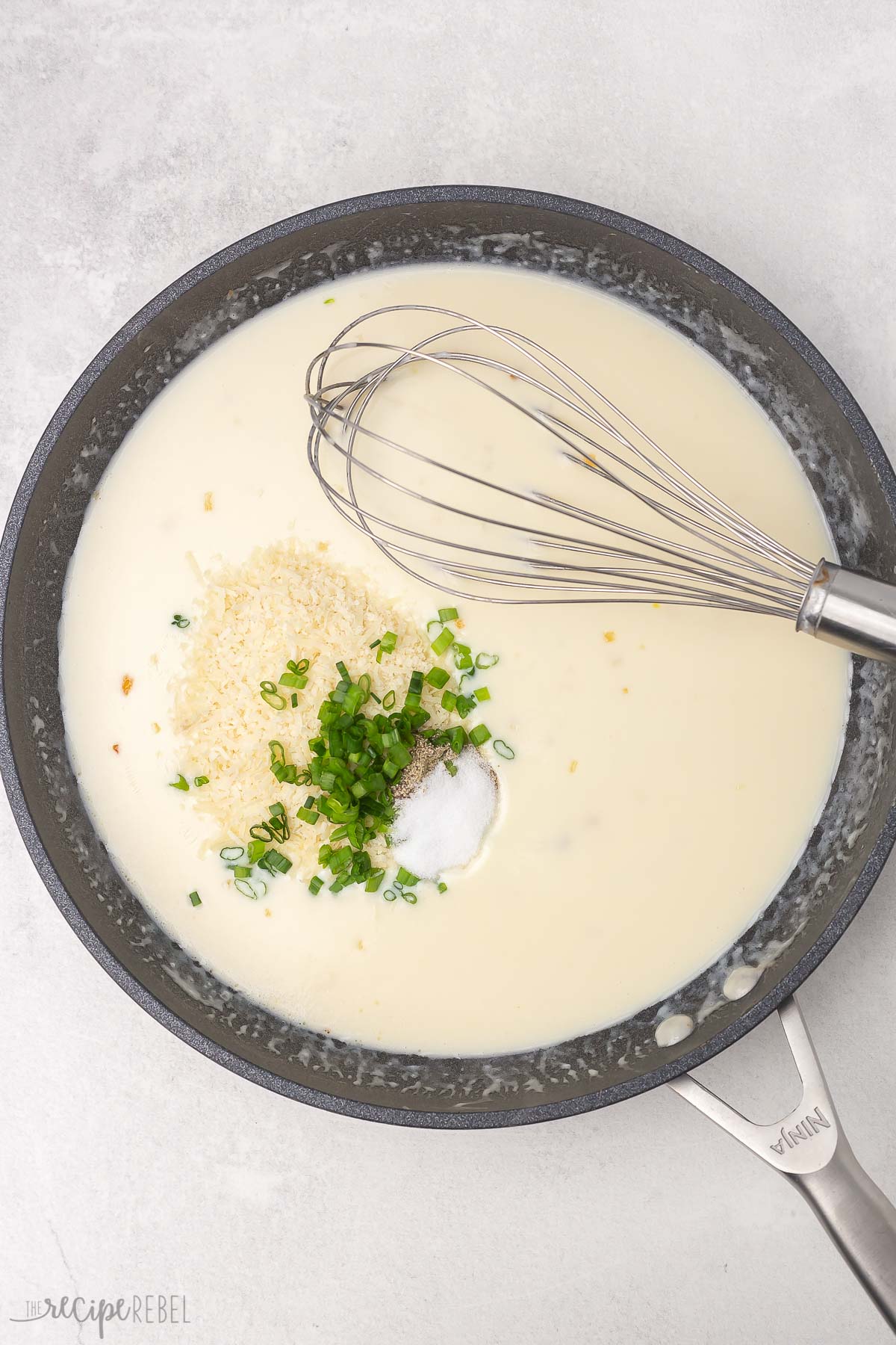 Top view of frying pan with a white creamy mixture in it with herbs and garlic and a whisk in it. 