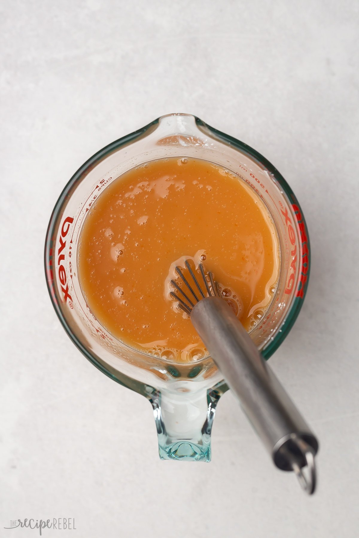 Top view of brown sweet and sour sauce in a glass jug with a whisk in it. 