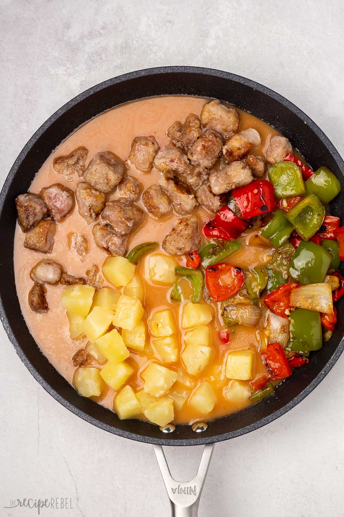 Top view of a pan with pork, sauteed veggies and pineapple chunks in it, as well as the sweet and sour sauce. 