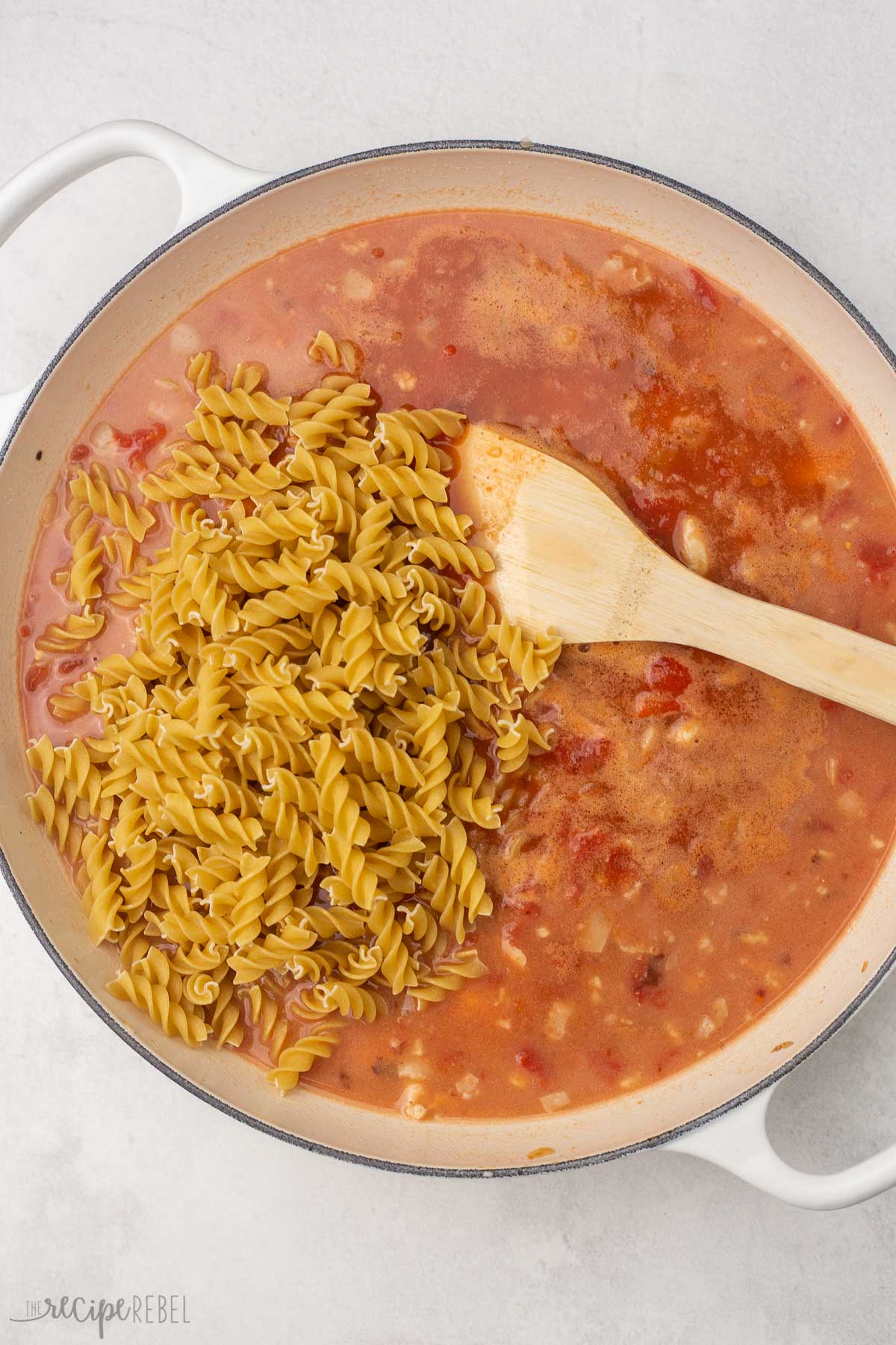 uncooked pasta lying on top of stirred ingredients in white dutch oven with wooden spatula.