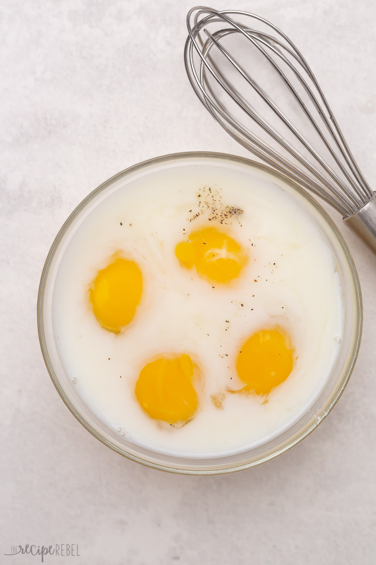 Top view of glass bowl with eggs, salt, pepper, and milk in it unmixed with a whisk beside it.