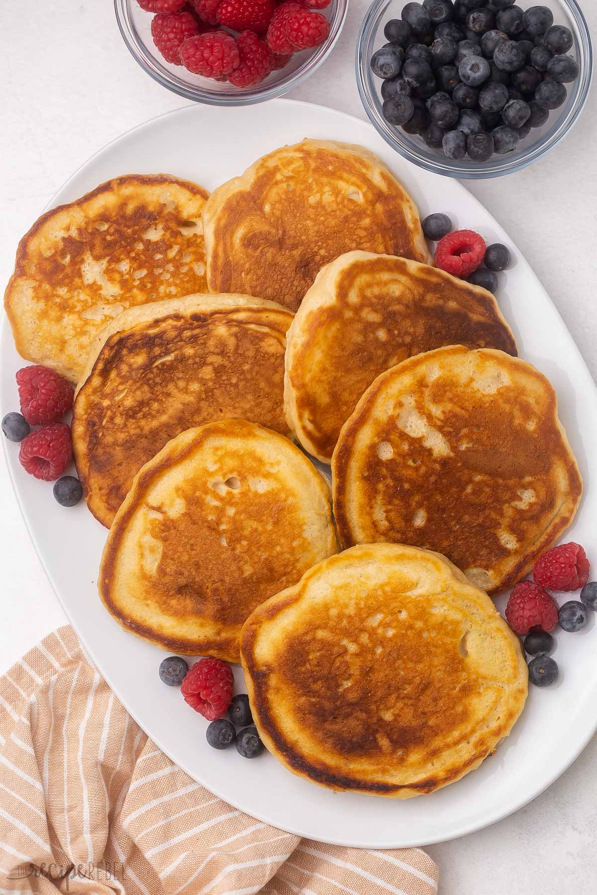 overhead view of white platter of pancakes with blueberries and raspberries spread around them.