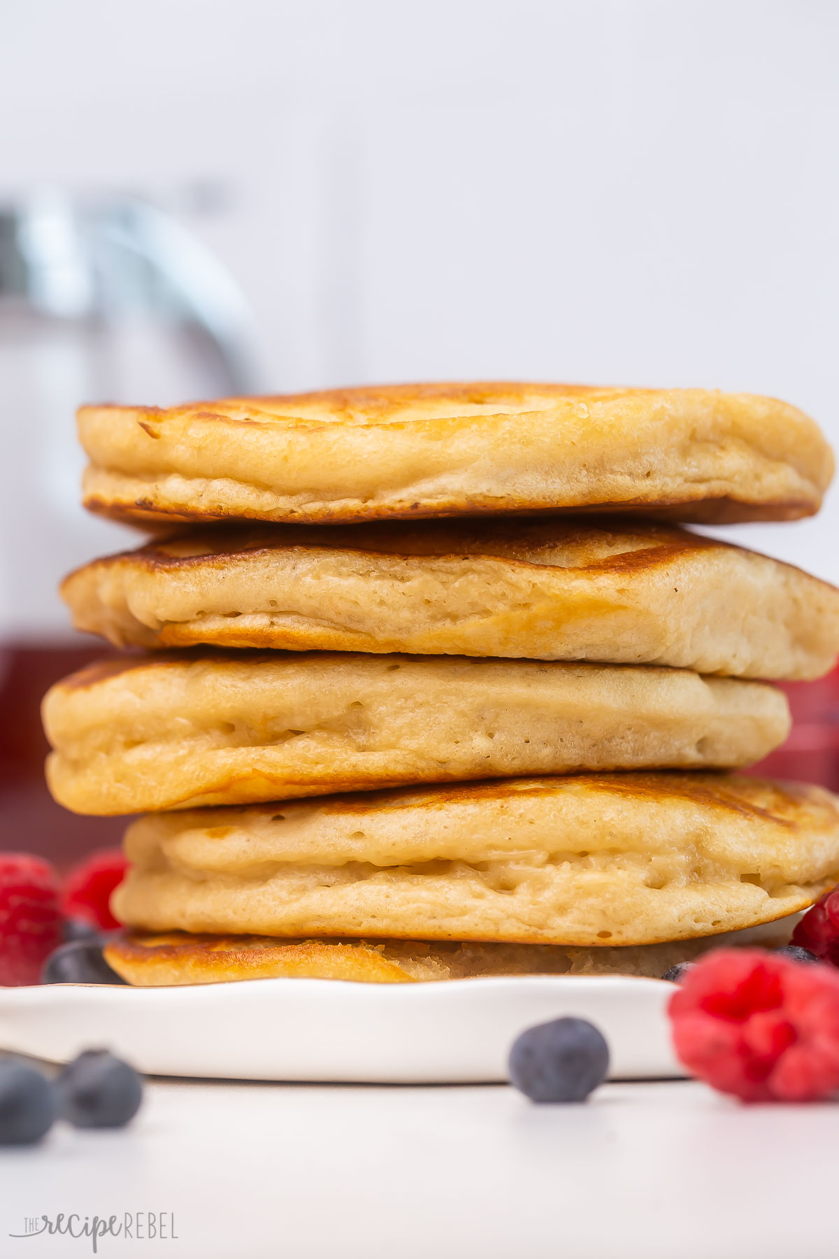 five buttermilk pancakes stacked on a white plate surrounded by berries.