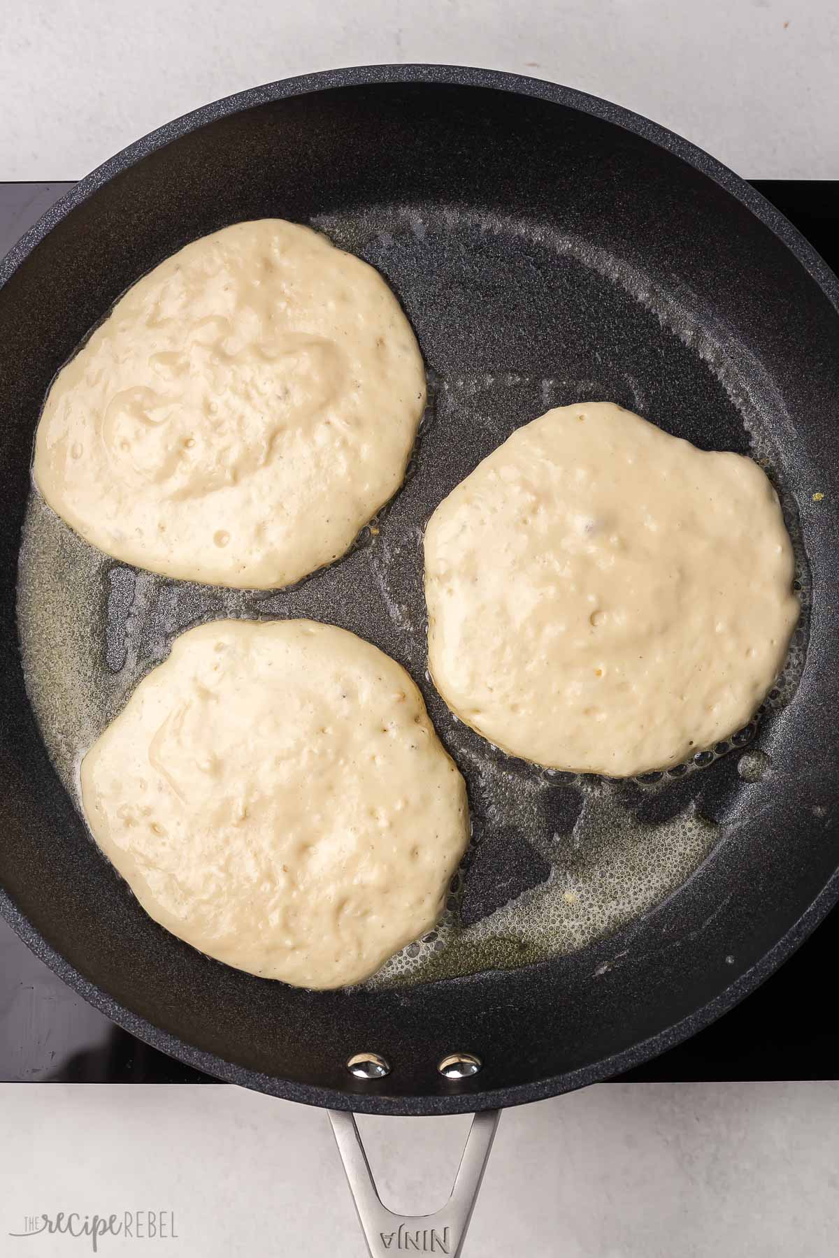 overhead view of black frying pan with three pancakes cooking inside.