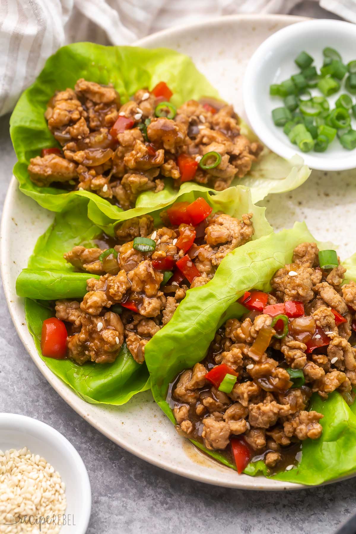 three chicken lettuce wraps lying on a plate with green onions and seasame seeds beside.