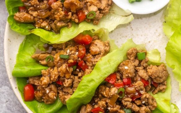 three chicken lettuce wraps lying on a plate with chopped green onions beside.