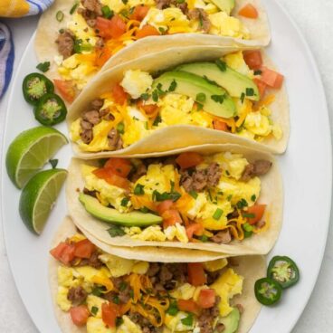 Top view of four breakfast tacos on a white platter with extra toppings placed beside.