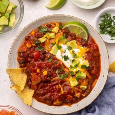 Top view of a white bowl of vegetarian chilli with toppings around it on the table.