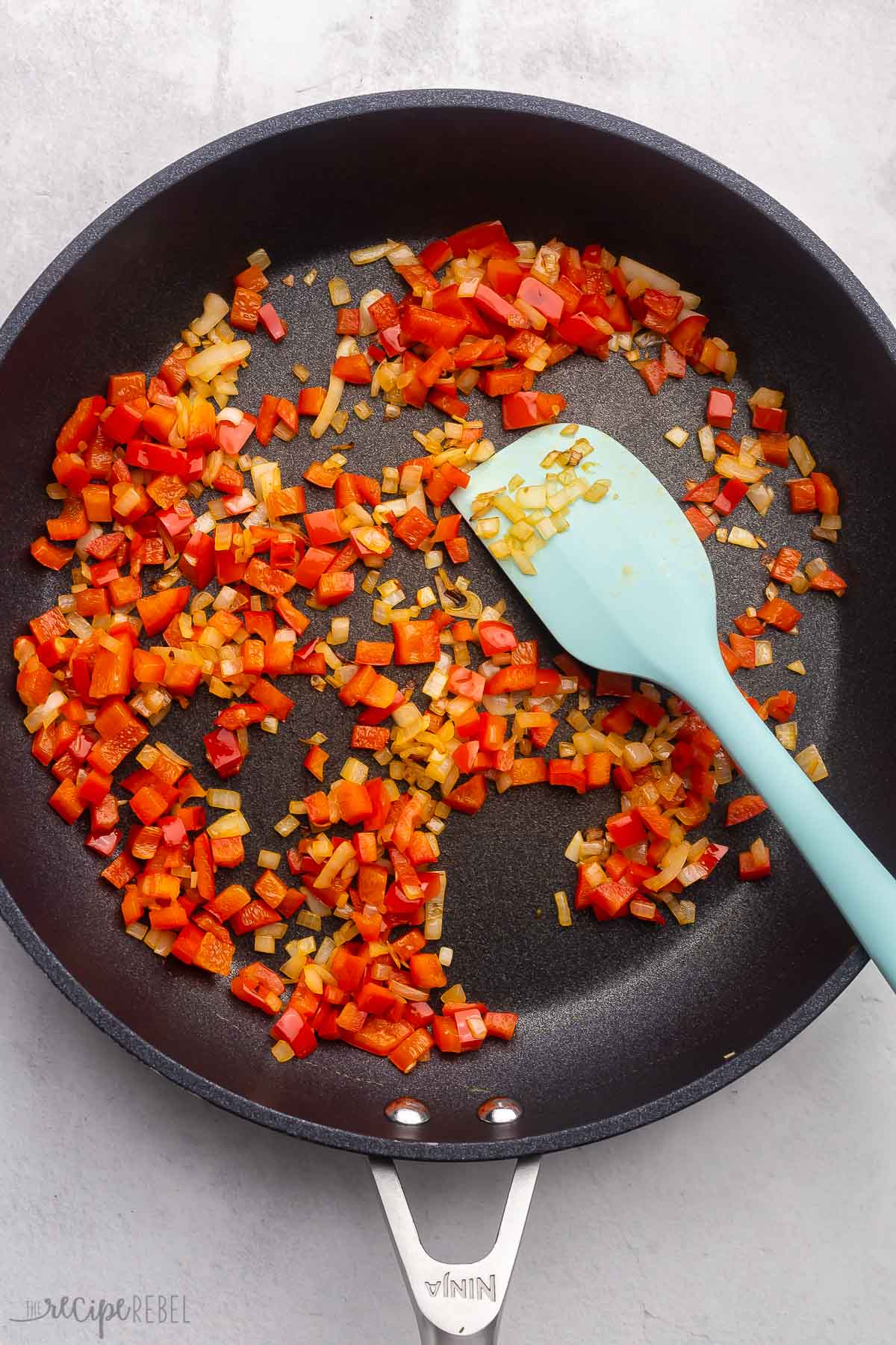 Top view of pan with chopped onions and red peppers being fried in it with a blue spatula in it. 