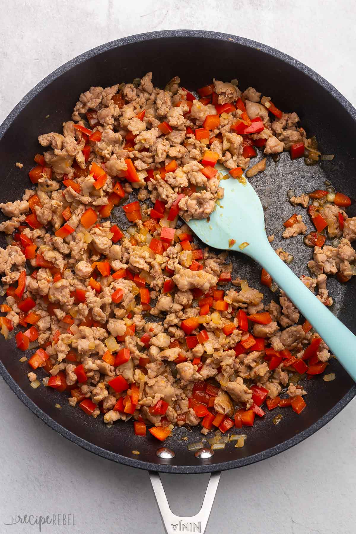Top view of pan with chopped onions and red peppers being cooked with browned chicken sausage and a spatula in the pan. 