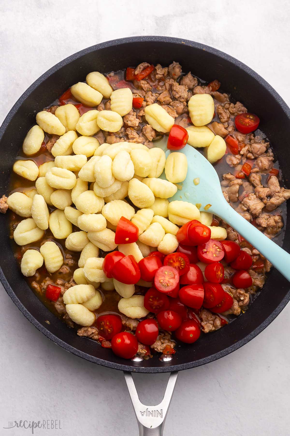 Top view of a black pan with sausage mix in the bottom, with gnocchi and chopped tomatoes on top and a spatula in the mix. 