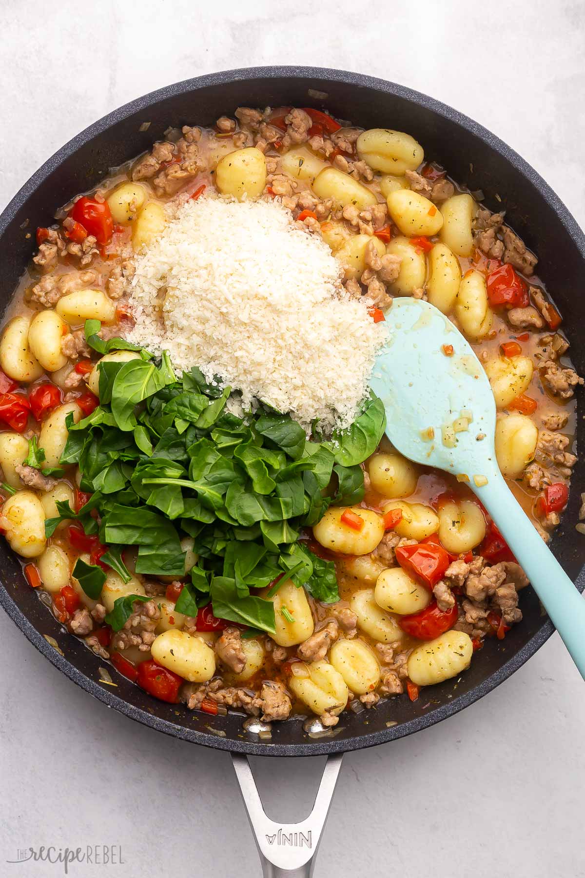 Top view of a black pan with a sausage mix, gnocchi, chopped tomatoes in the bottom, and a spatula in the mix, with grated parmesan cheese and spinach leaves on top.