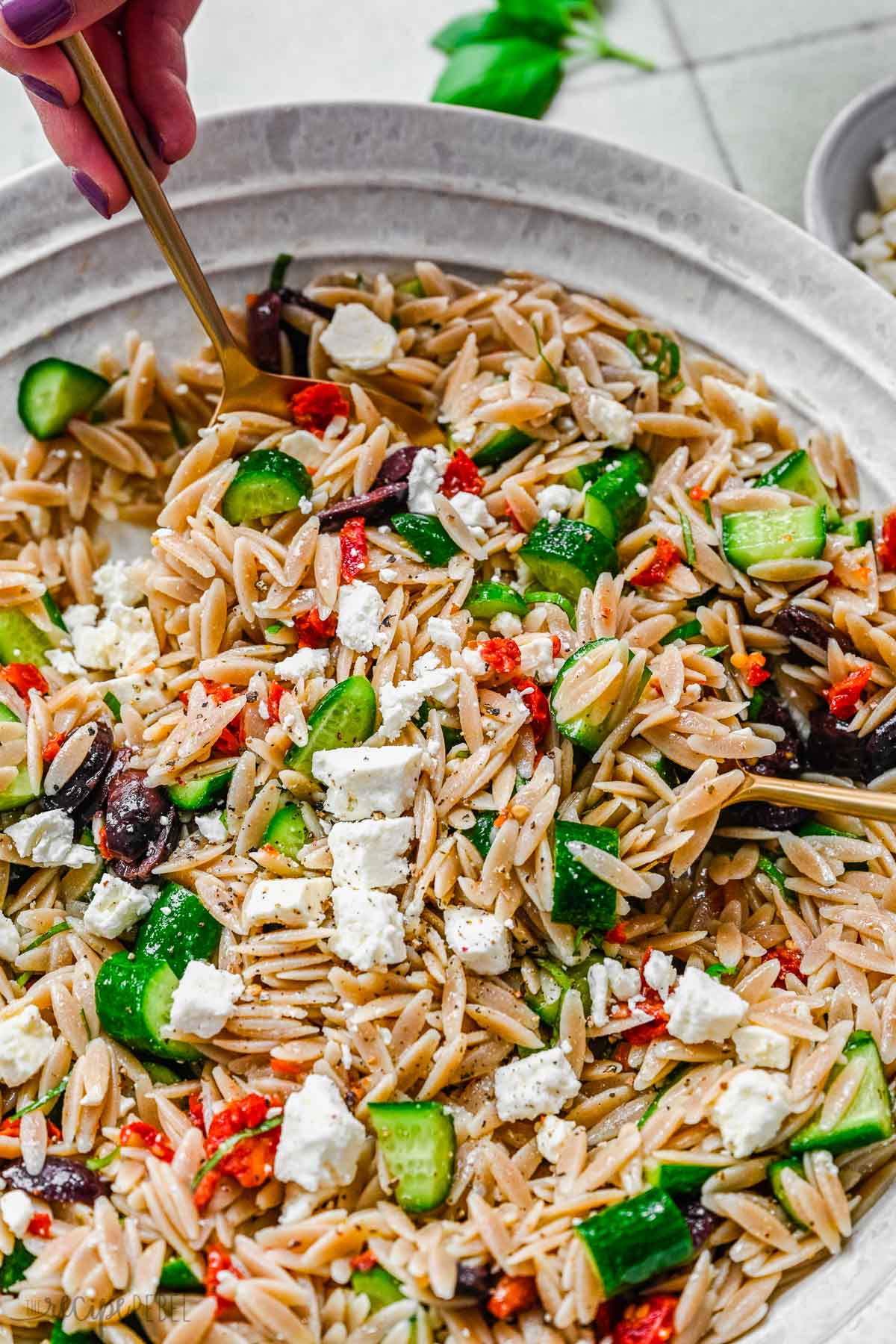 Top view of Greek Pasta Salad in a pasta bowl with someone holding a gold spoon in it. 