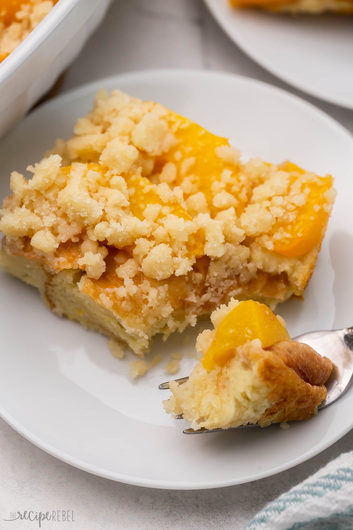 Close up of a slice of peach cobbler French toast on a white plate with a fork having taken a forkful of it. 