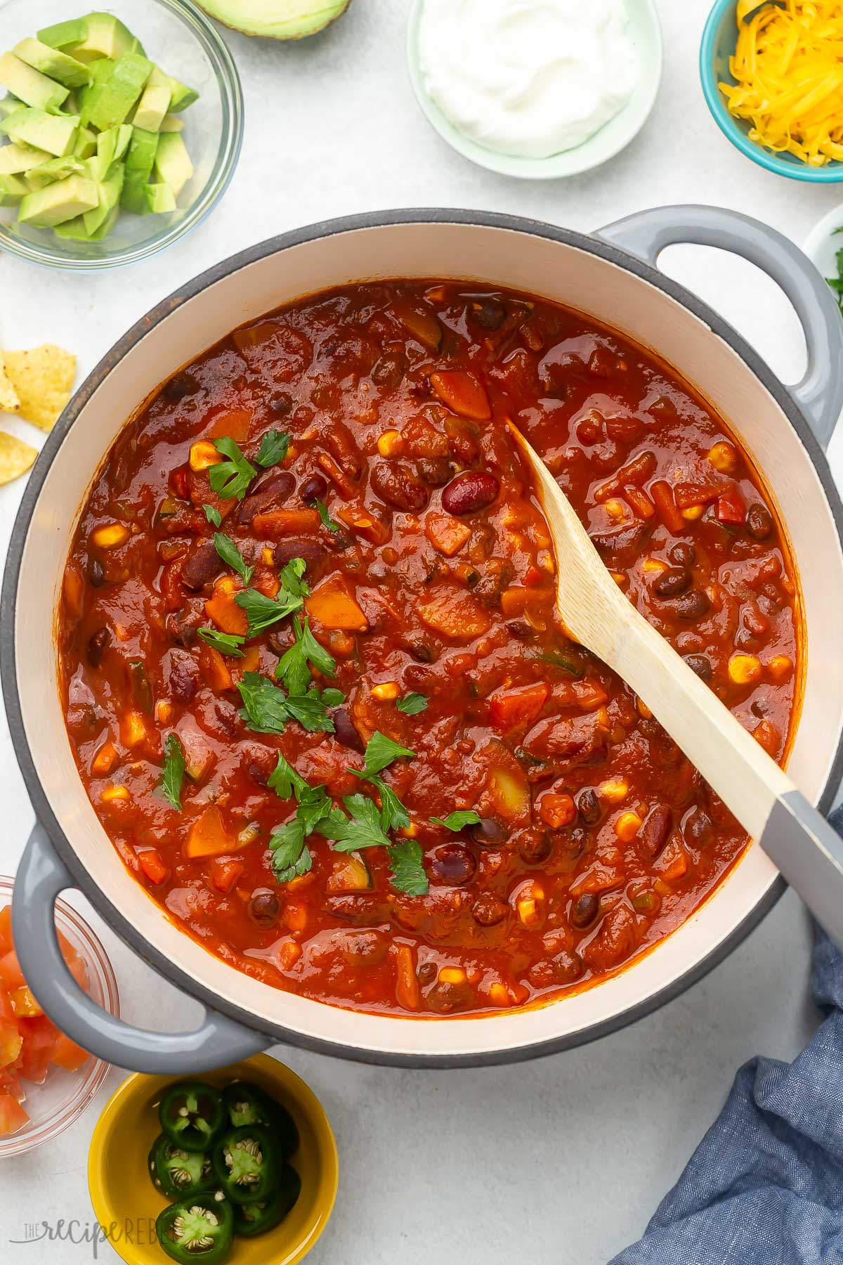 Overhead view of a large pot of vegetarian chilli with wooden ladle and toppings.
