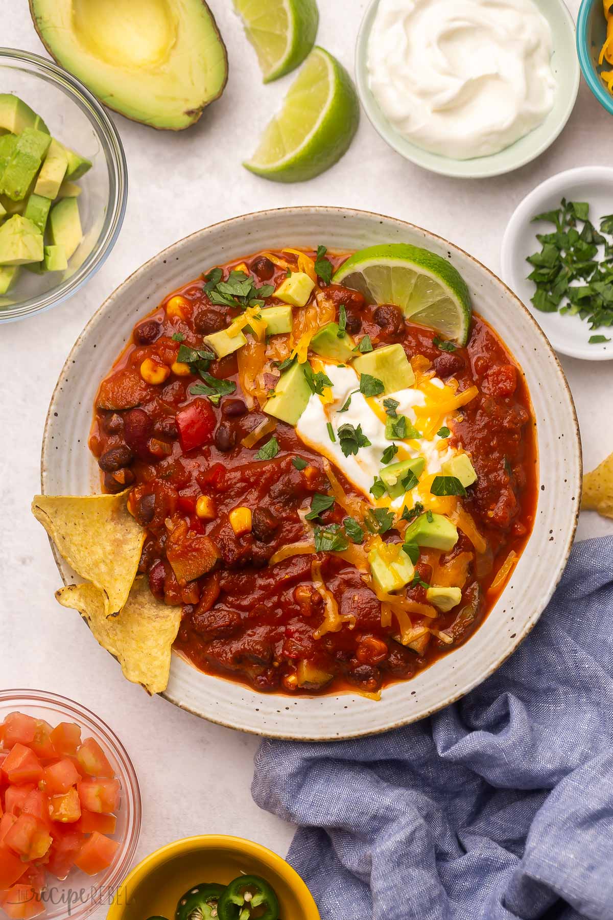 Top view of a white bowl of vegetarian chilli with toppings around it on the table.