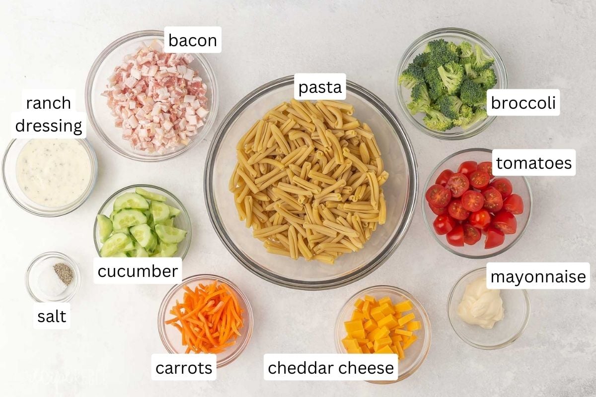 Top view of ingredients needed to make Bacon Ranch Pasta Salad in small bowls on a white table with labels over them. 