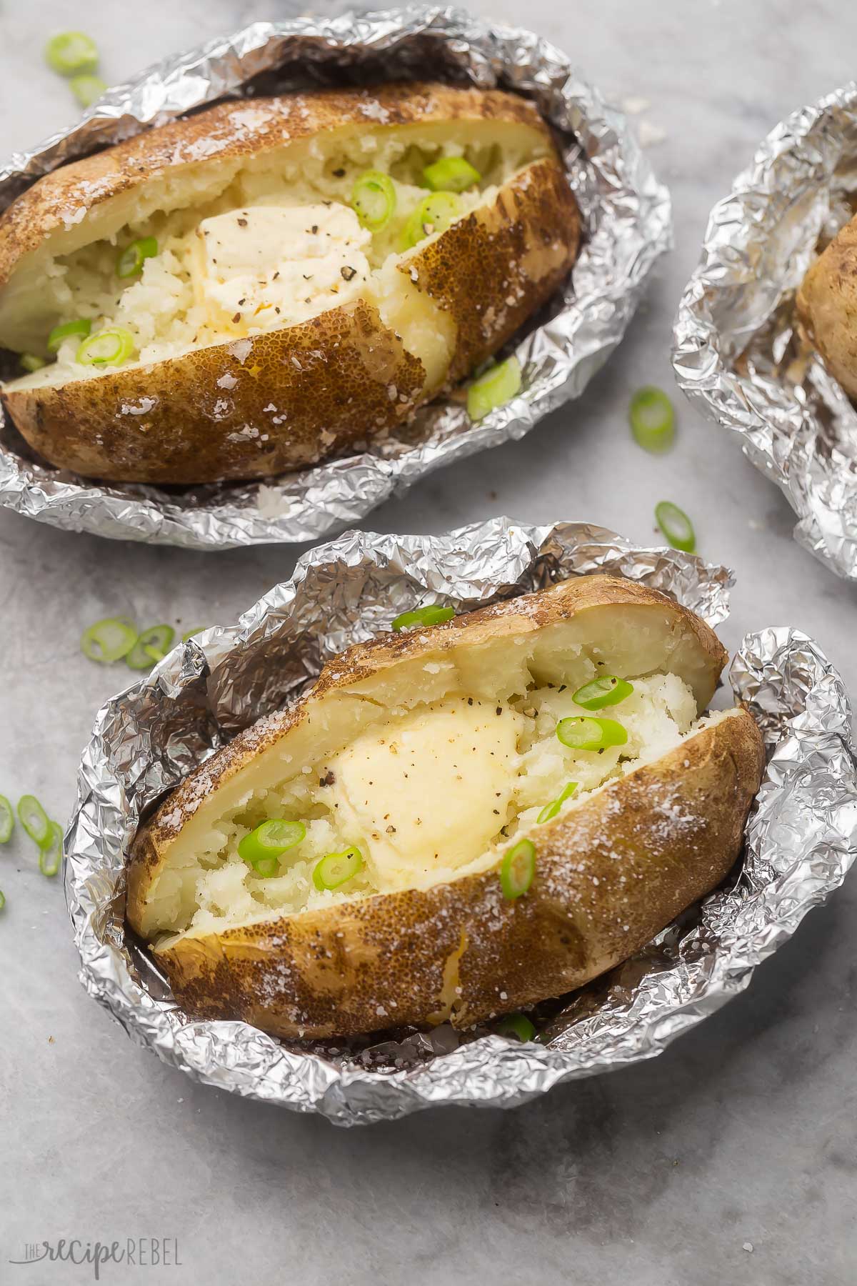 Top view of two baked potatoes unwrapped from foil packets, with butter melting inside them, and some chopped green onions on top. 