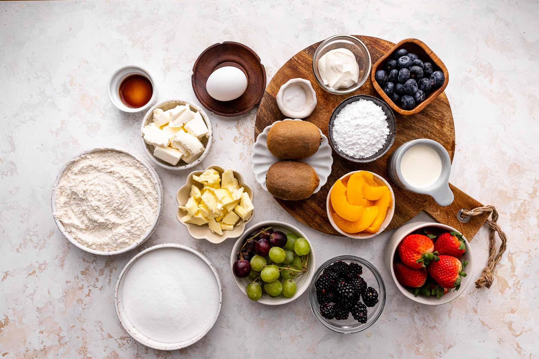 Top view of ingredients needed to make fuit pizz, in small bowls on a gray surface. 