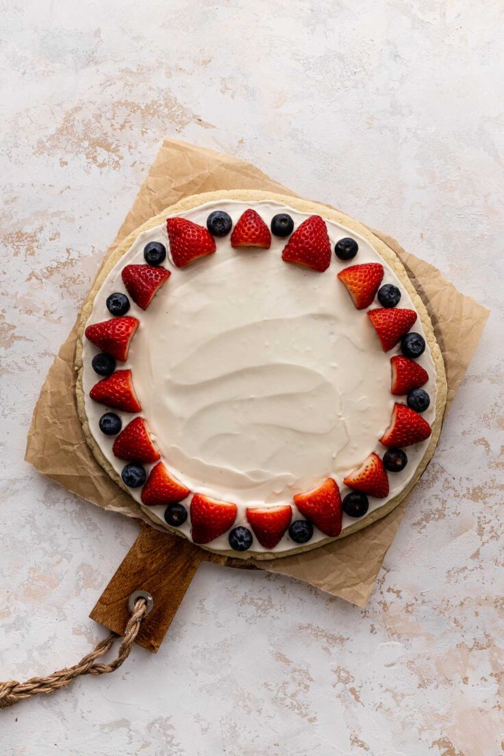 Top view of sugar cookie base with cream cheese mixture on top and a row of blueberries and strawberries near the edge of the pizza circle. 