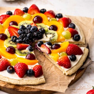 Closse up of a fruit pizza on a chopping board with a slice cut out of it.