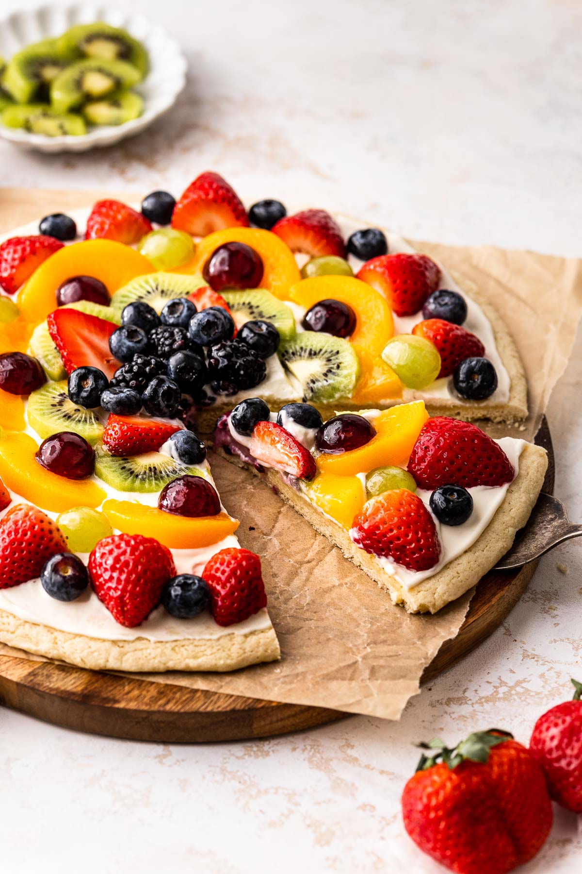 Closse up of a fruit pizza on a chopping board with a slice cut out of it. 