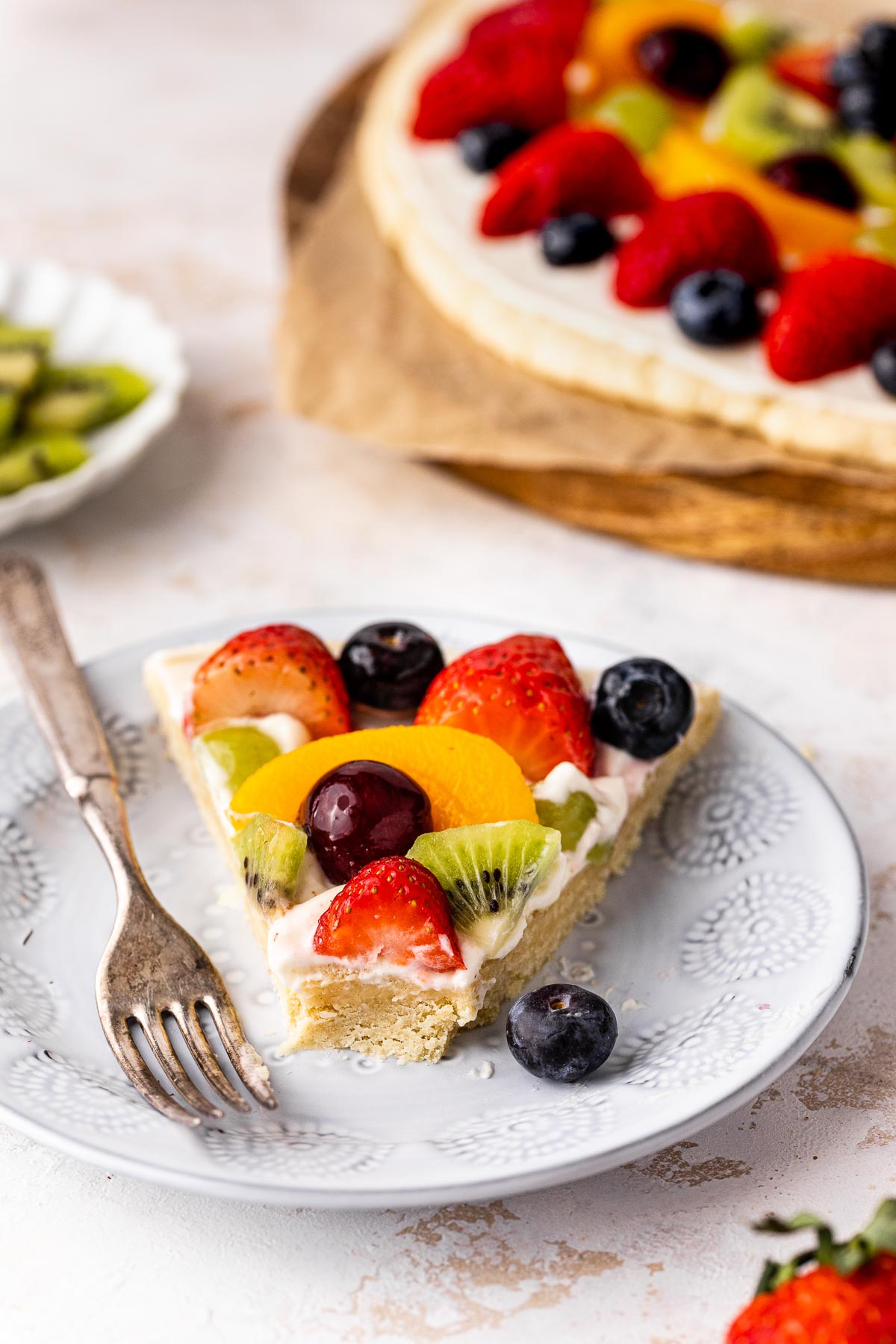 Close up of a slice of fruit pizza on a white plate with a fork on the side.