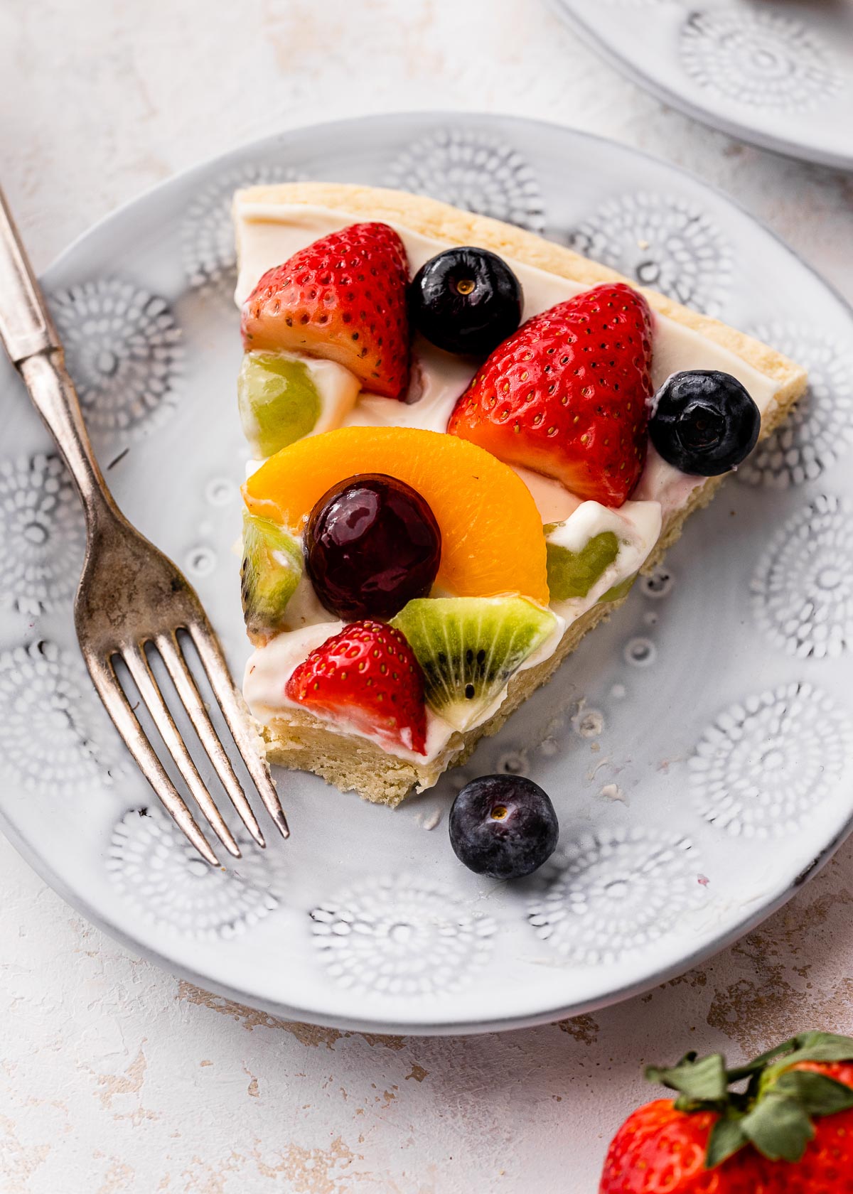 Top view of a slice of fruit pizza on a white plate with a fork on the side.