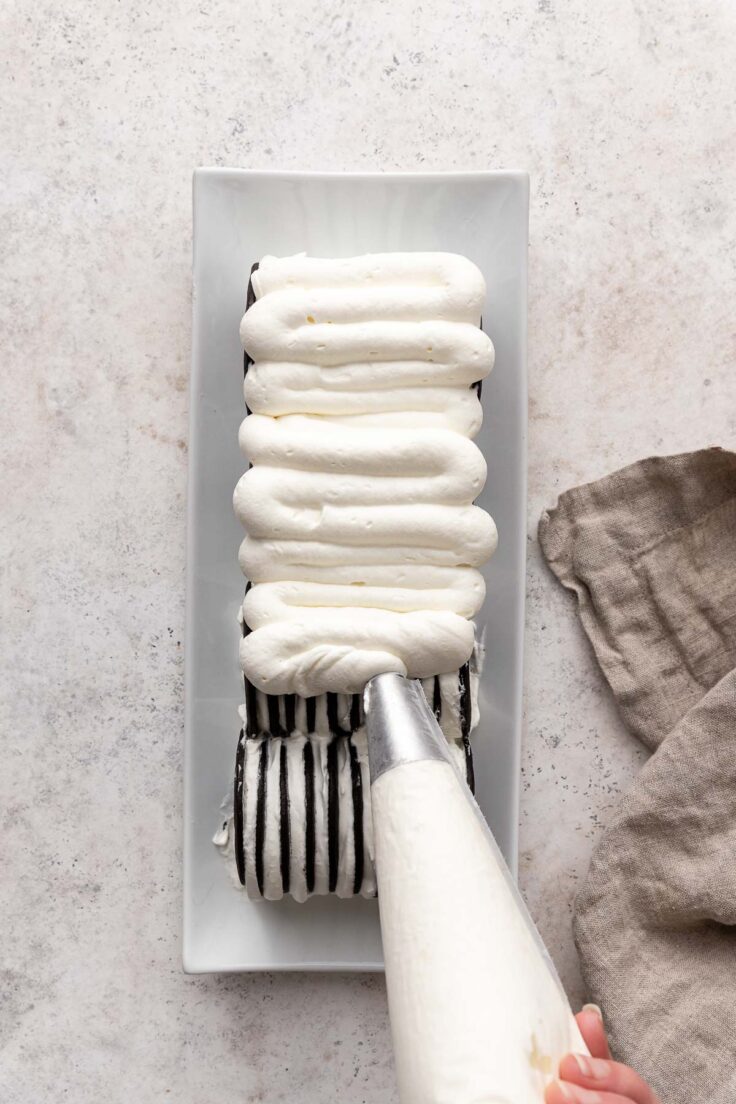 Top view of a block of chocolate ice box cake being topped with whipping cream mixture with a piping bag. . 