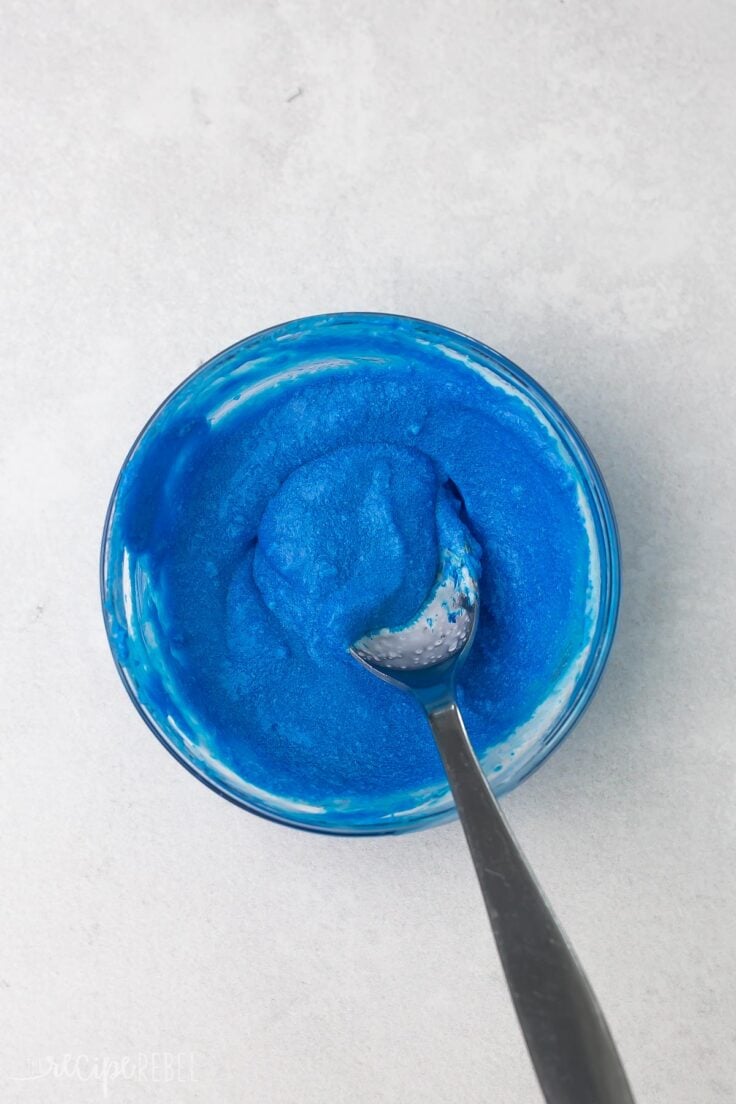 Top view of a small glass mixing bowl with a blue liquid in it being stirred with a spoon. 