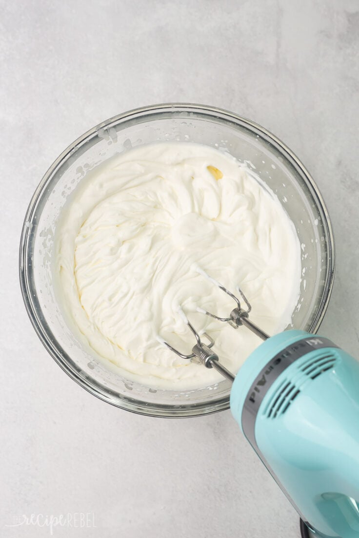 Top view of a glass mixing bowl with a creamy white mixture in it being whisked with a hand mixer. 