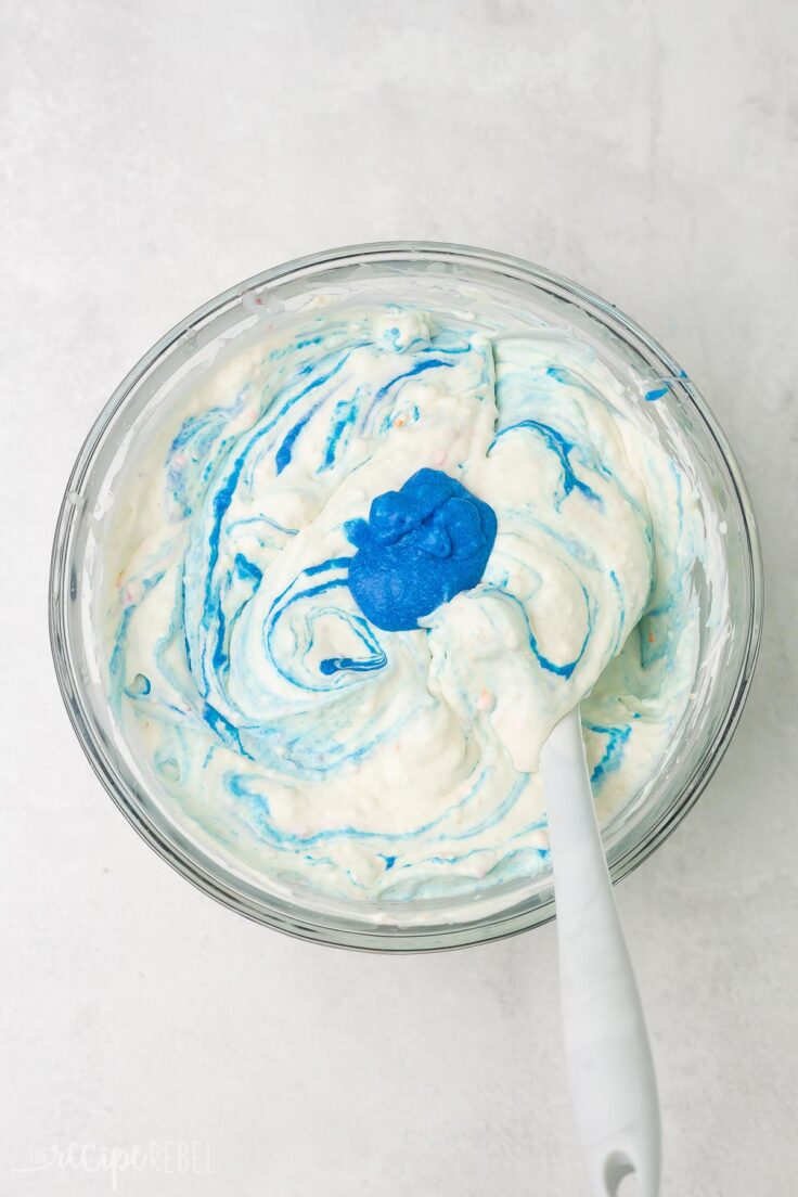 Top view of a glass mixing bowl filled with ice cream mix with a blue liquid being mixed into it. 