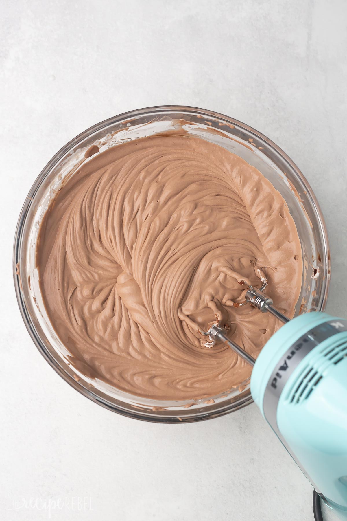 Top view of a glass mixing bowl with a chocolate ice cream mixture being whisked with a hand mixer. 