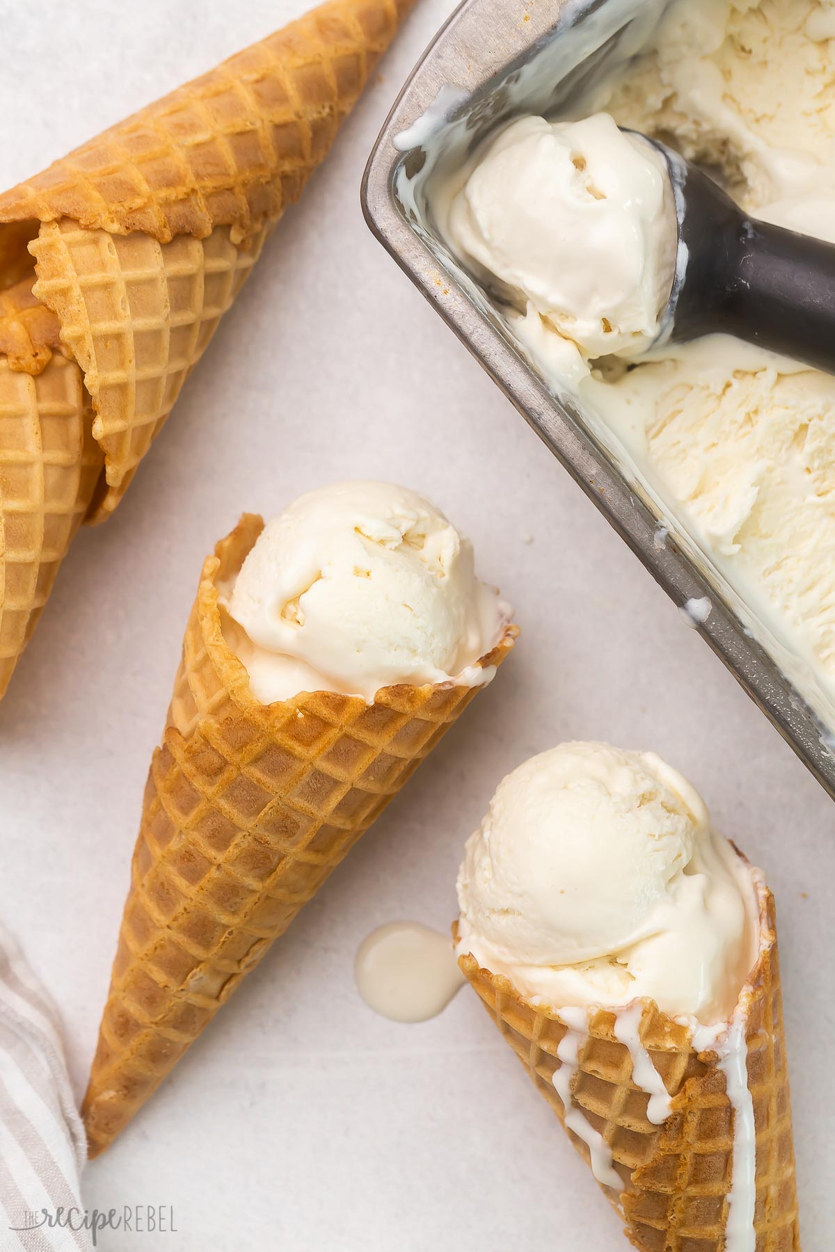 Top view of waffle cones with vanilla ice cream in it lying on worktop next to a tub of vanilla ice cream. 