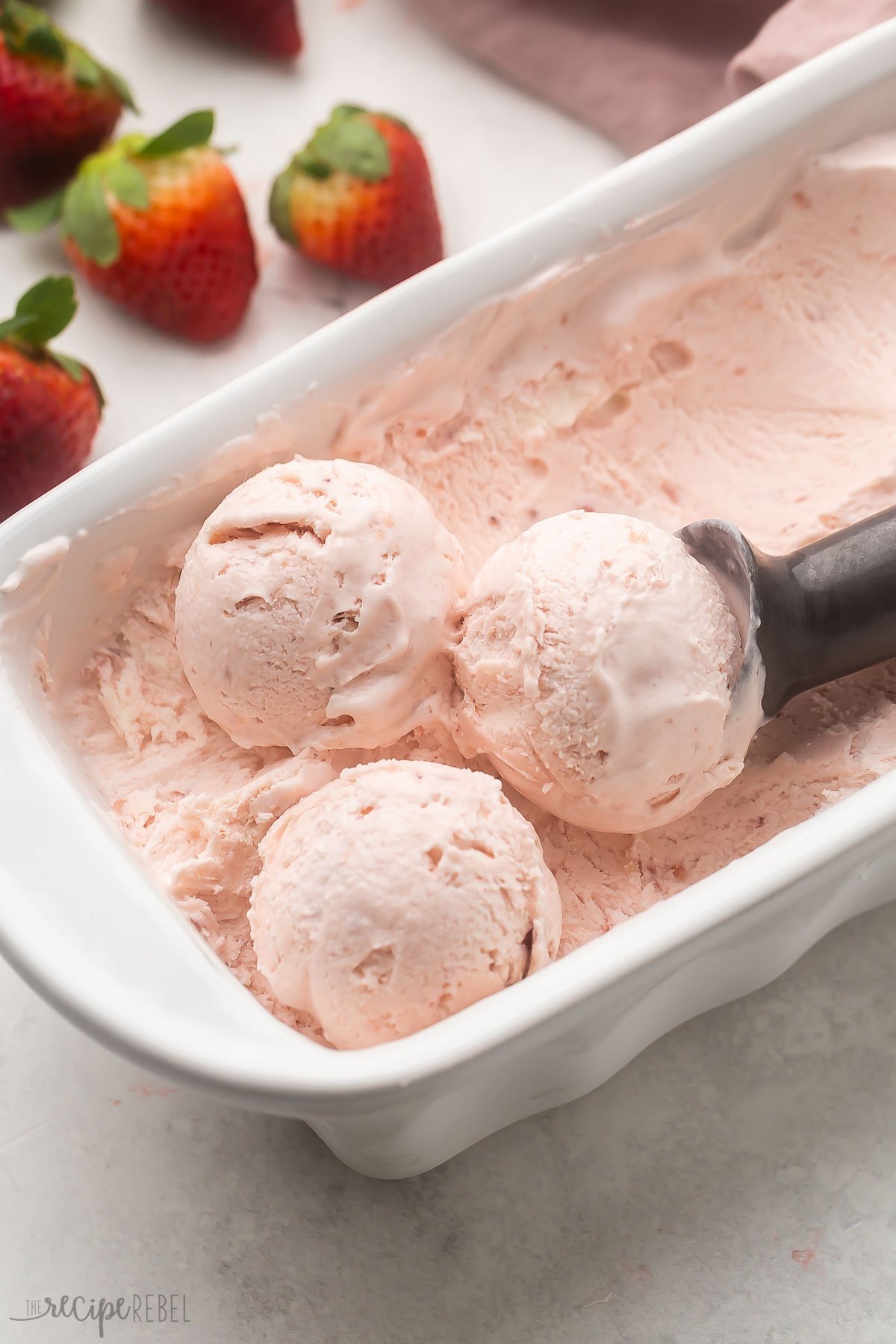 White dish filled with strawberry ice cream in it, being scooped out by an ice cream scoop. 