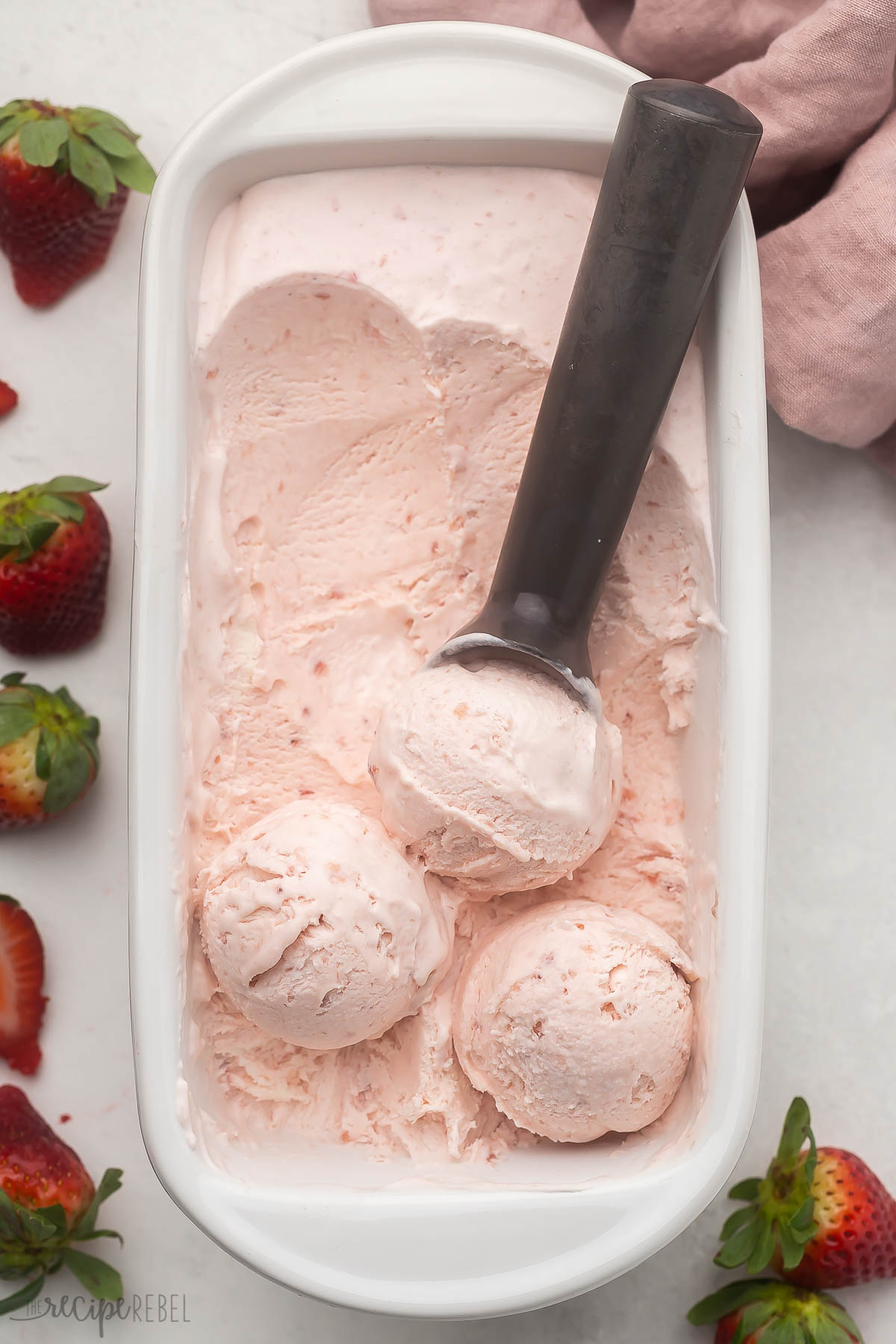 Top view of a white dish filled with strawberry ice cream in it, being scooped out by an ice cream scoop.