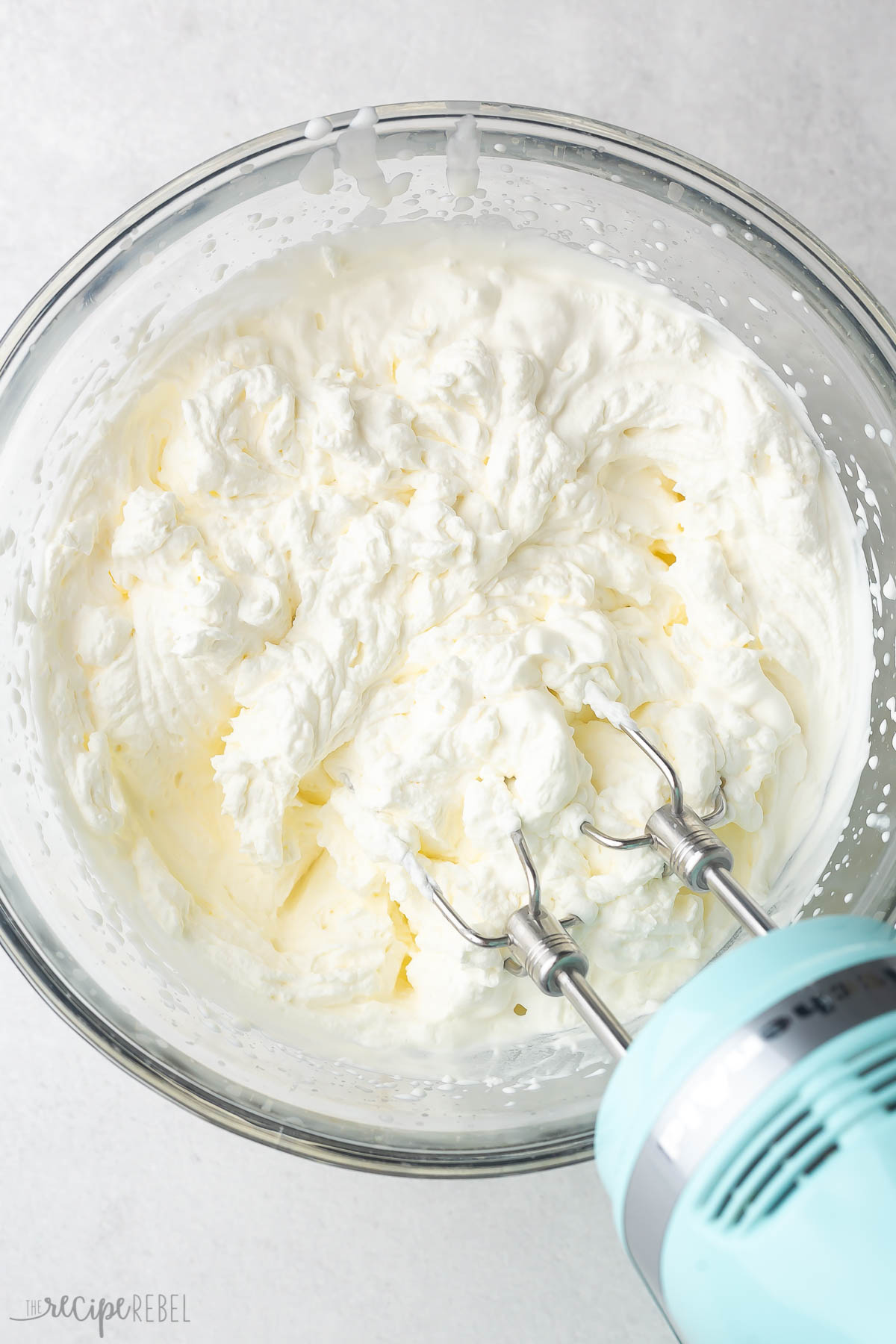 Top view of glass mixing bowl with heavy cream being whipped by an electric hand mixer.