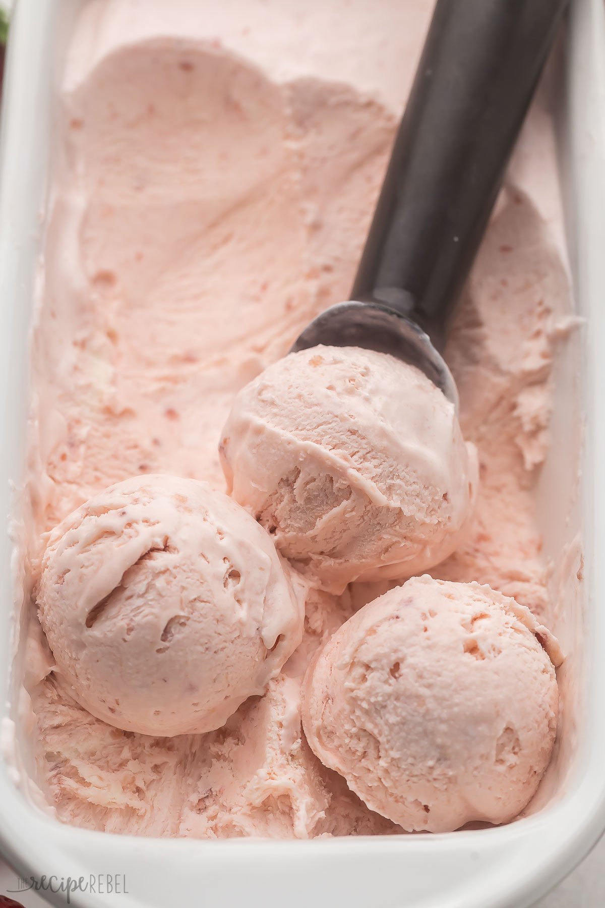 Top view close up of a white dish filled with strawberry ice cream in it, being scooped out by an ice cream scoop.