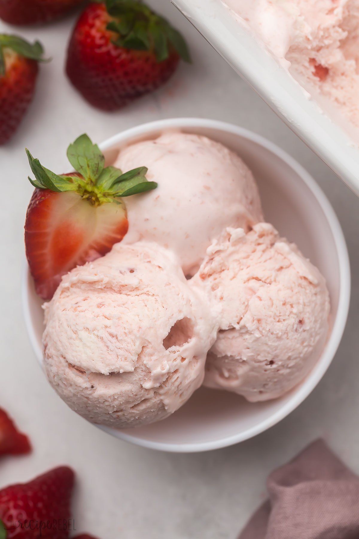 Top view of a white bowl with three scoops of strawberry ice cream in it and a slice of fresh strawberry.